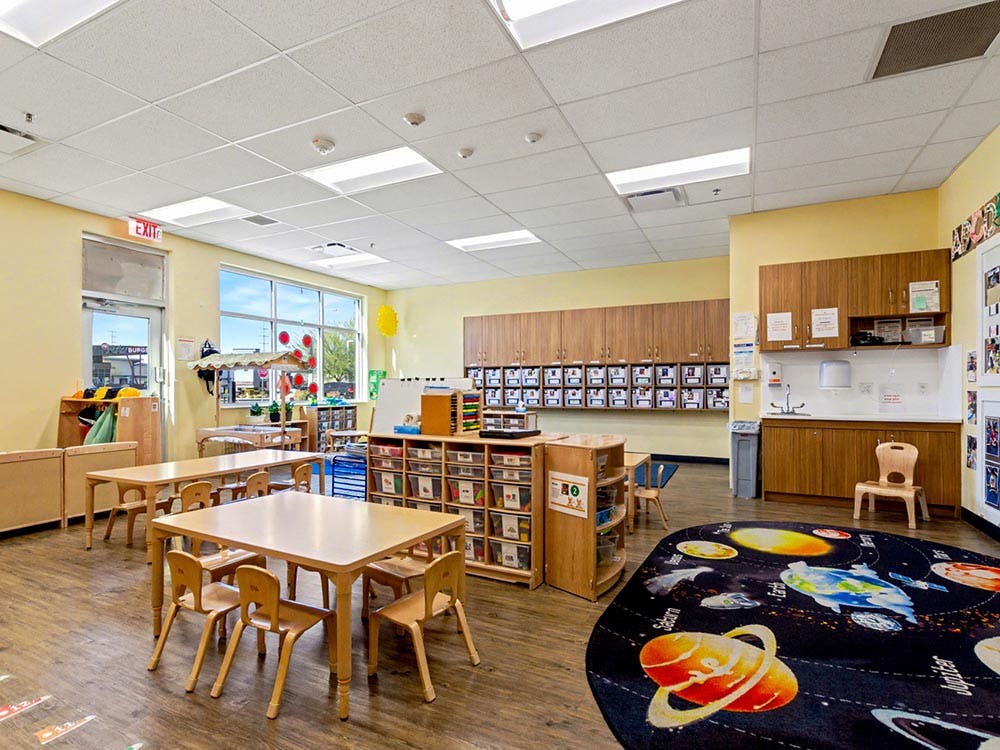 Classroom with small wooden tables and chairs, storage cabinets, and a planetary rug