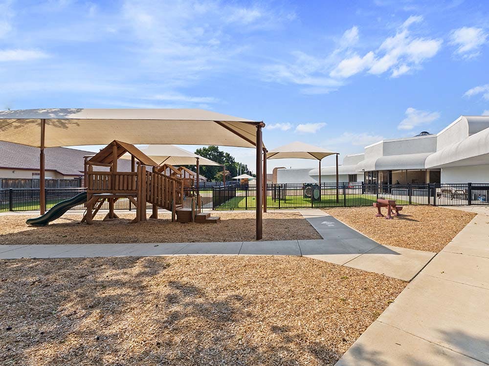 Outdoor playground with wood chips under a shaded canopy next to a building