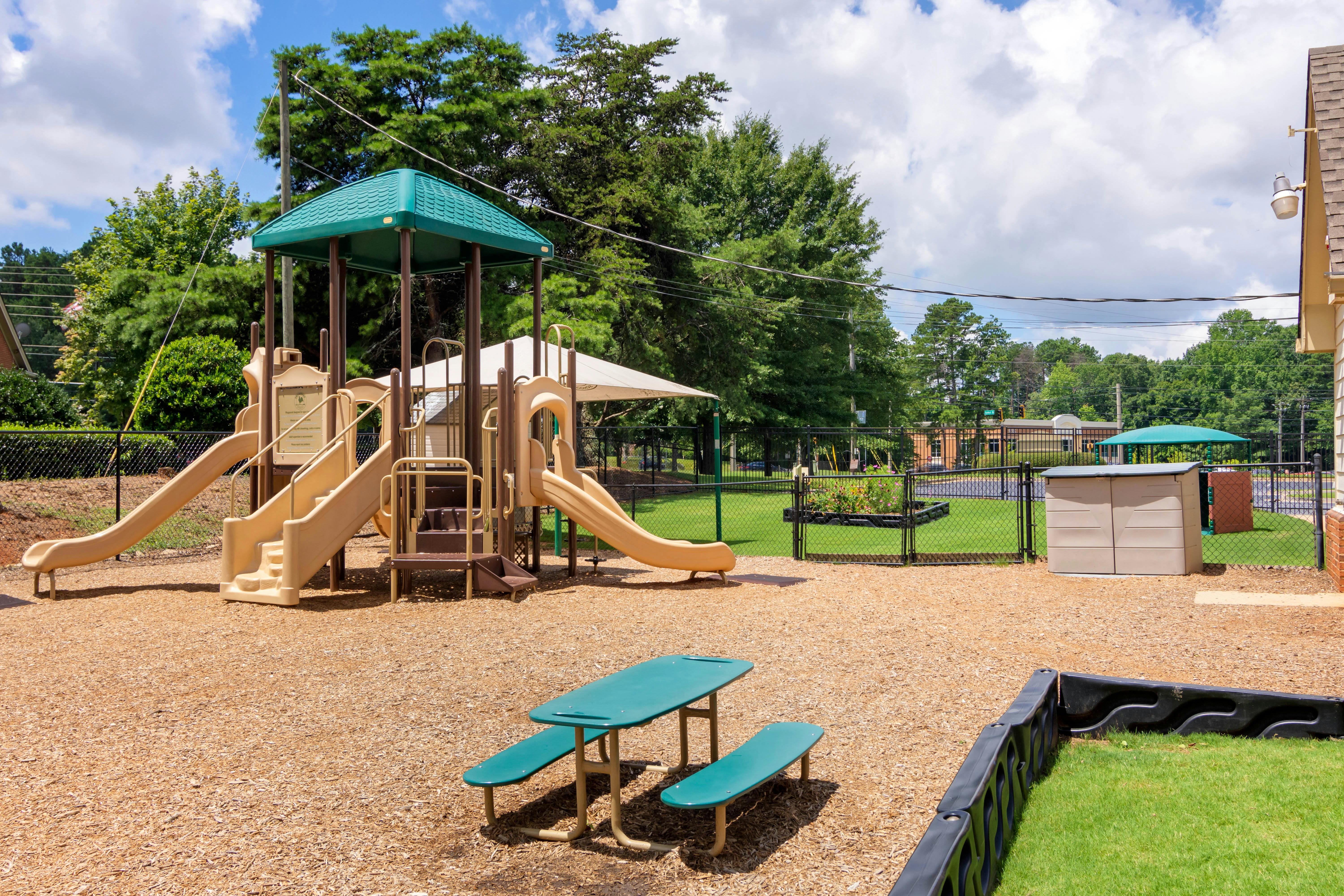 Playground with slides and stairs on wood chips, a green picnic table, and fenced grass area