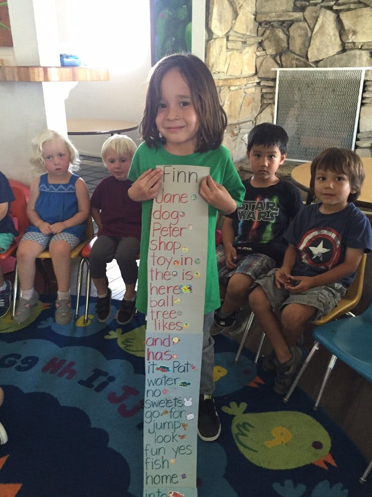 Child holding a long paper with words written on it, with other children seated behind