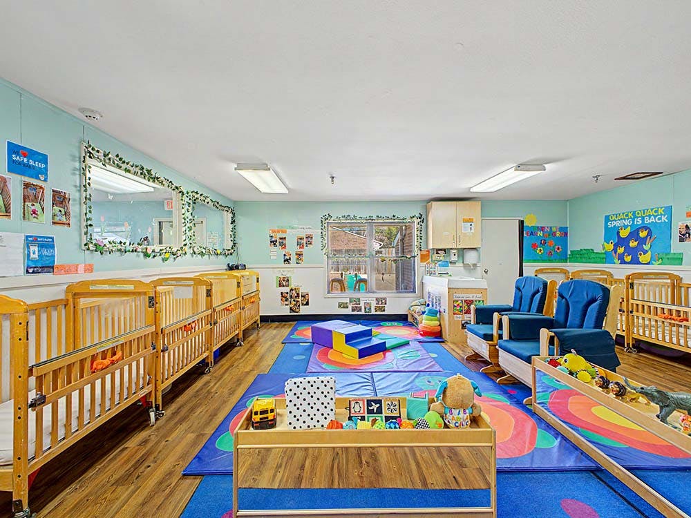 A colorful childcare classroom with several cribs, play mats, and rocking chairs
