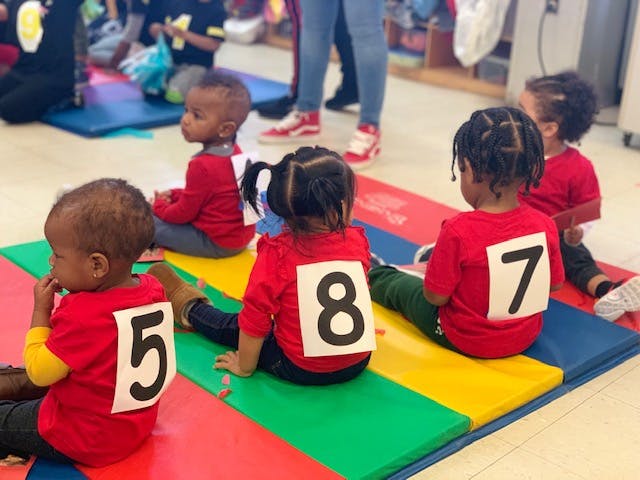 Children in red shirts with numbers 5, 8, and 7 on their backs sitting on mats