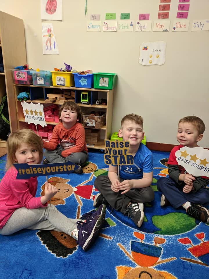 Four children sitting on a rug holding signs that say "Raise Your Star"