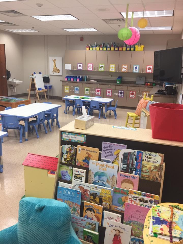 Classroom with small tables and chairs, a bookshelf with children's books, and storage cabinets