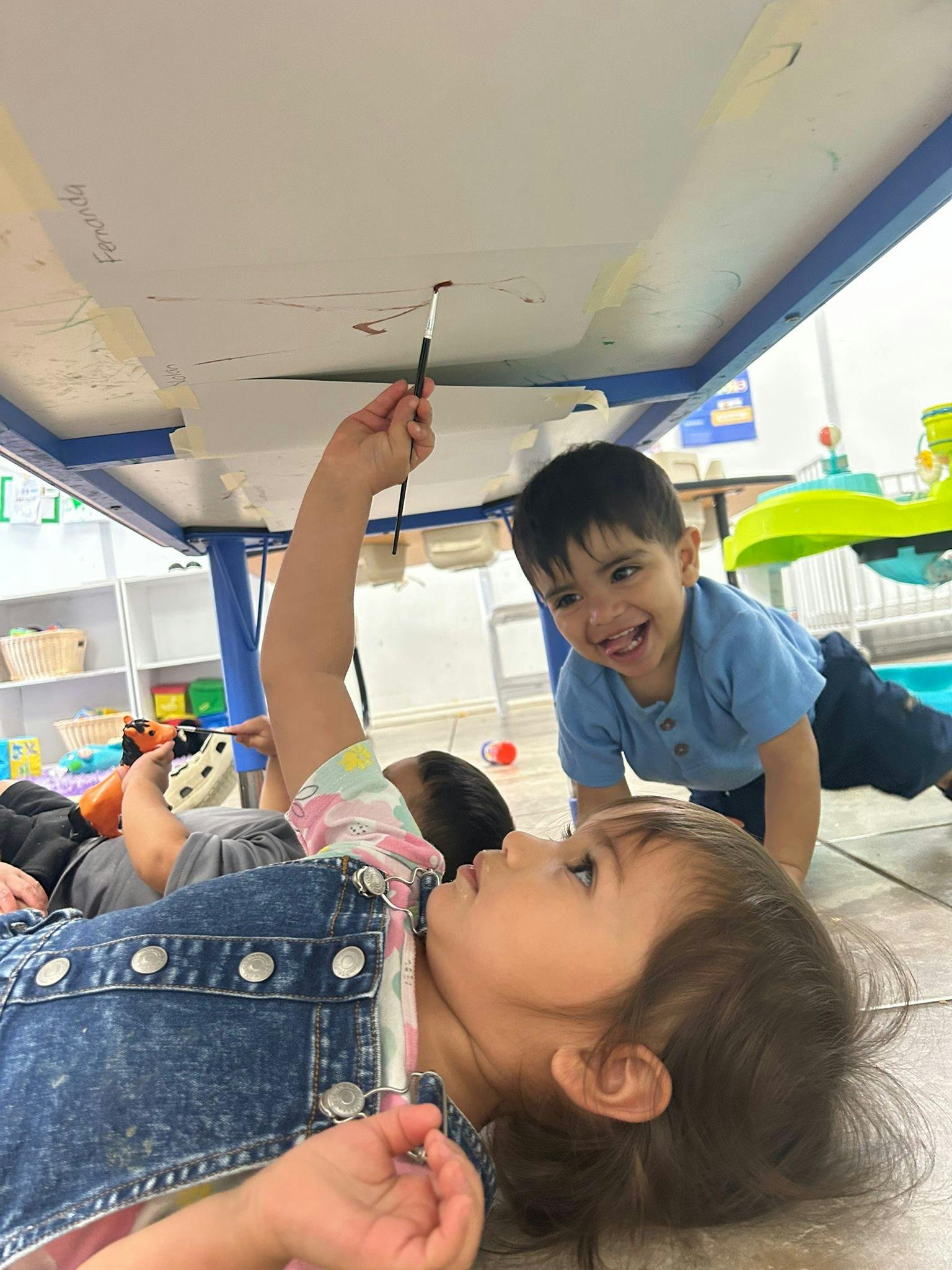 Child lying on back painting under a table, another child watching