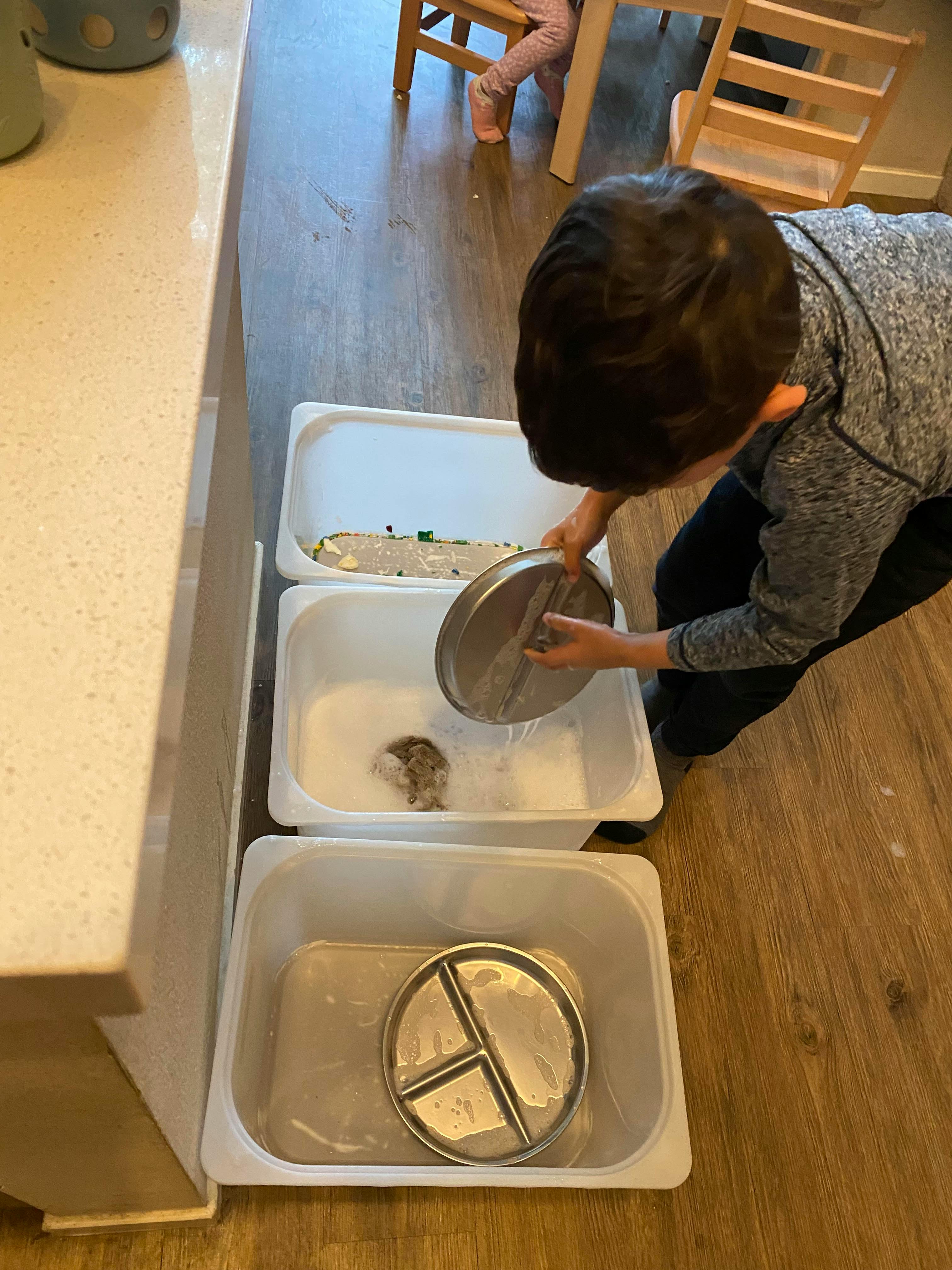 Child washing metal plates in plastic tubs filled with soapy water