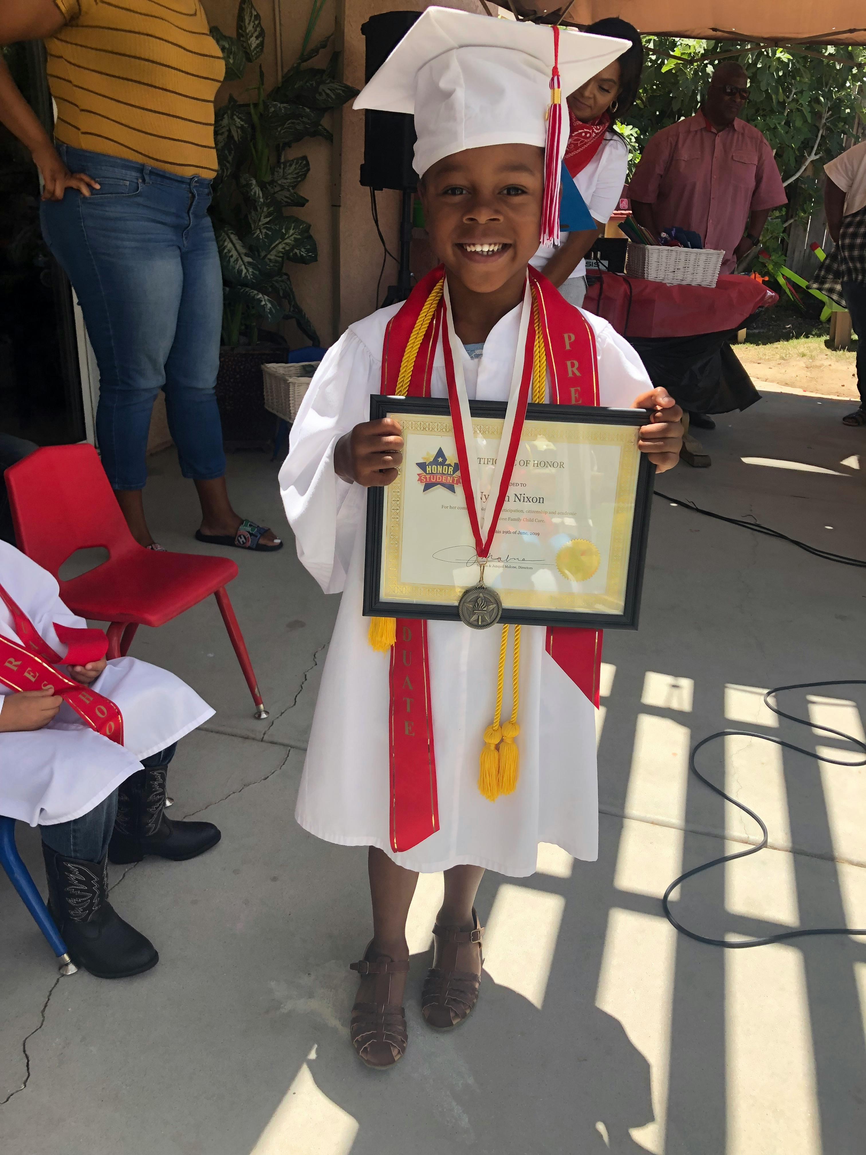 Child in graduation cap and gown holding a framed certificate and matted certificate