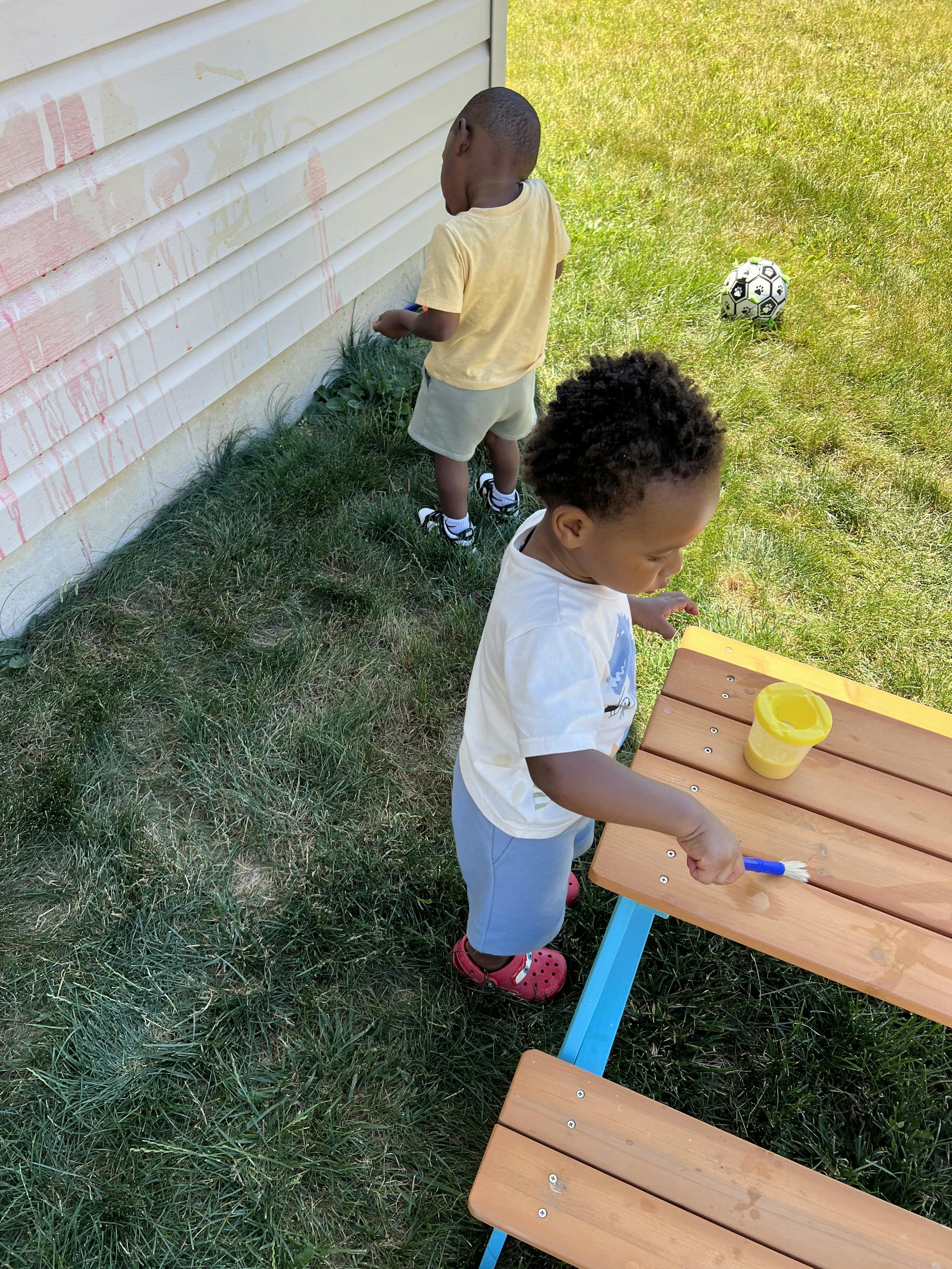Two children playing with paint outside near a wall and a small wooden table