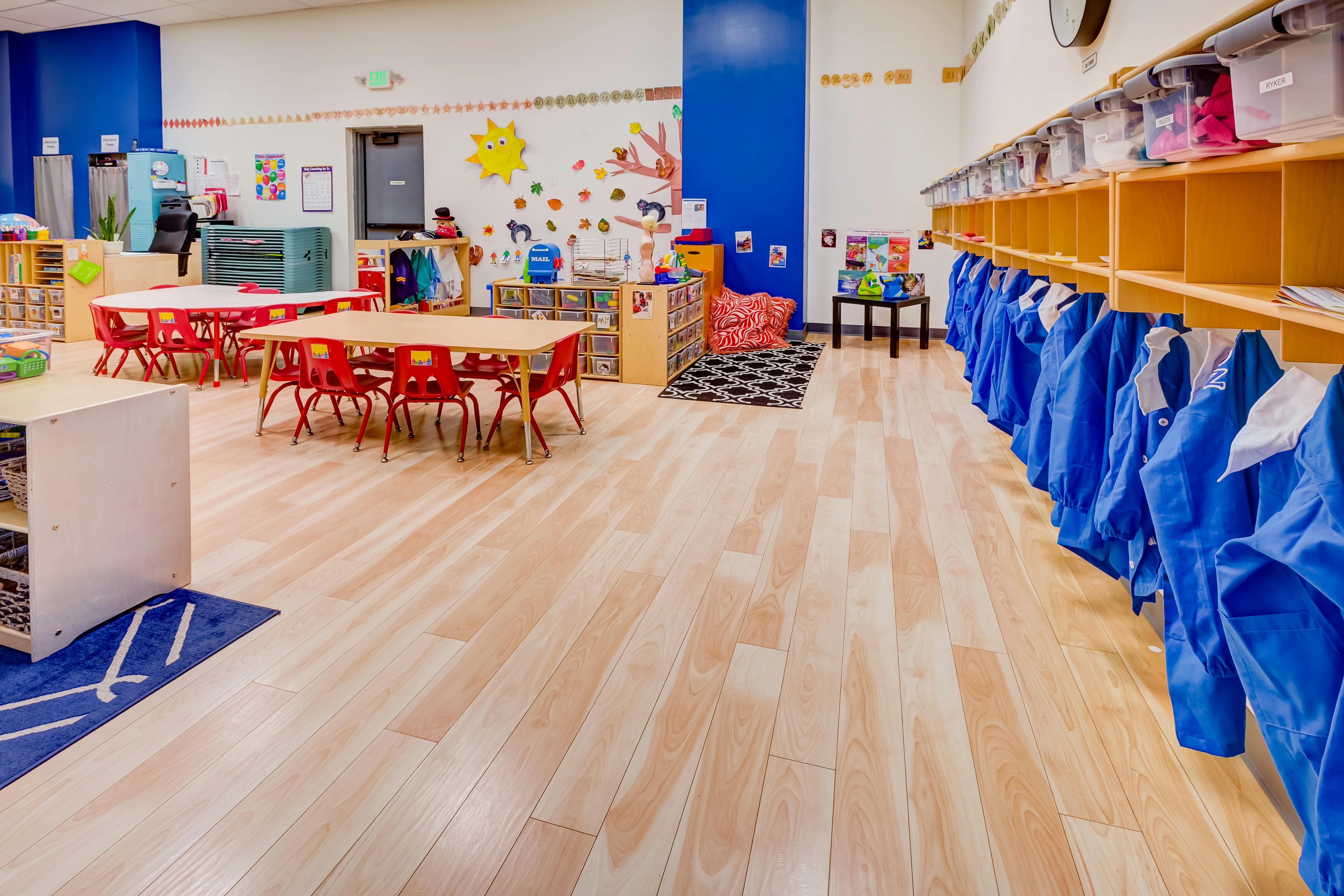 Empty classroom with small tables and chairs, cubbies with blue smocks
