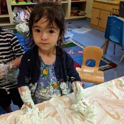 Child with hands covered in white foam on a table in a classroom
