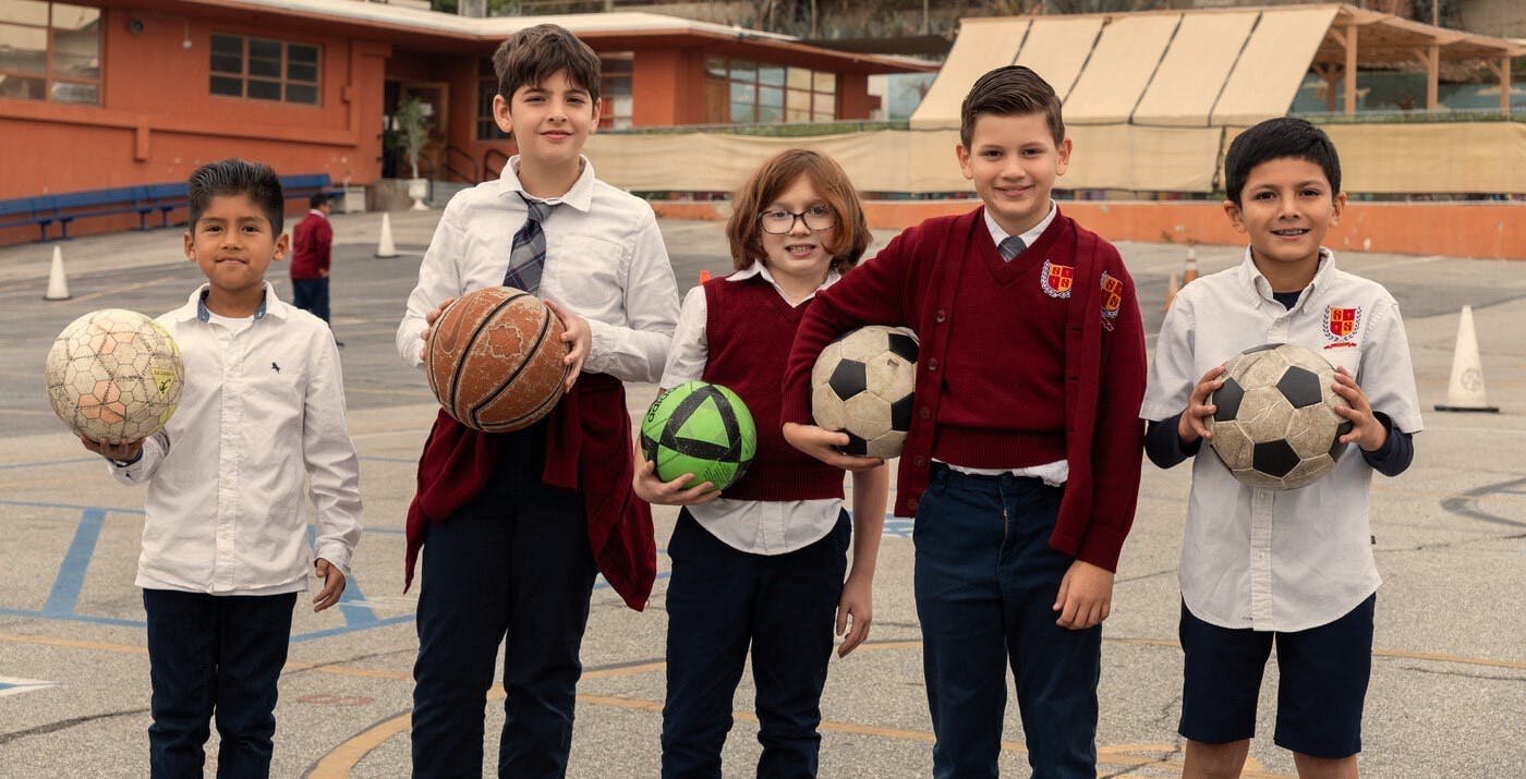 Five children in school uniforms holding soccer balls and a basketball on a playground