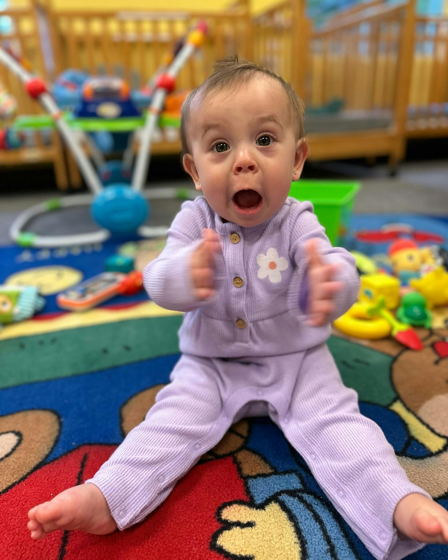 Child in purple outfit sitting on a colorful rug with mouth open