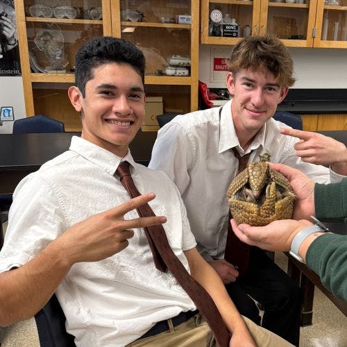 Two teens with a person holding an armadillo in a classroom
