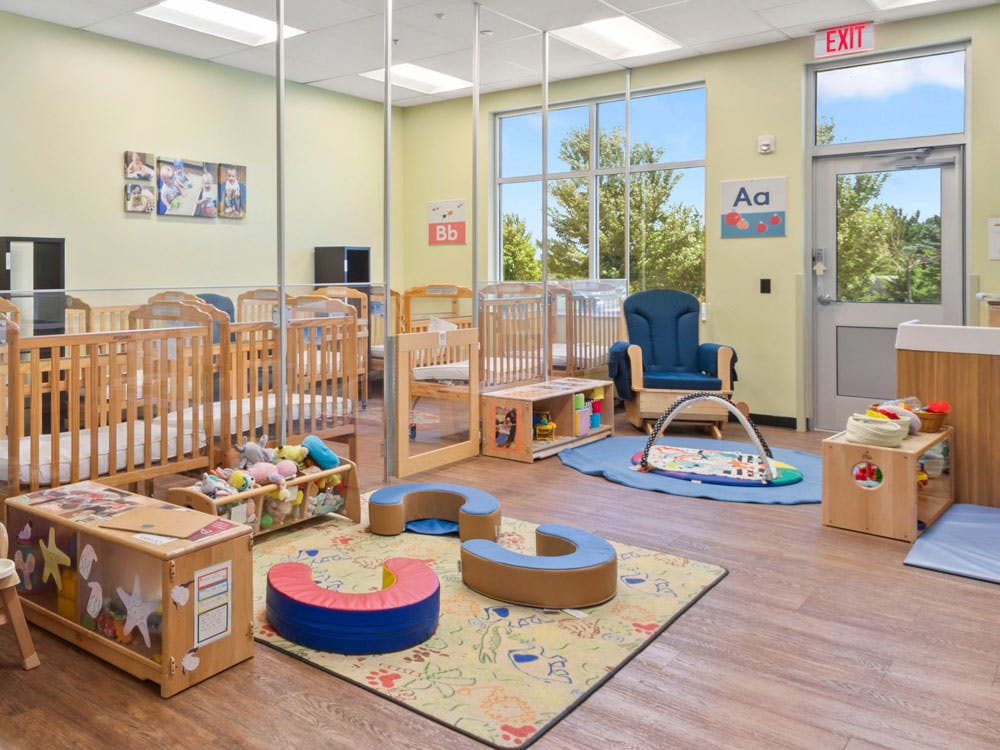 A brightly lit childcare room with multiple cribs, a play mat, and small furniture