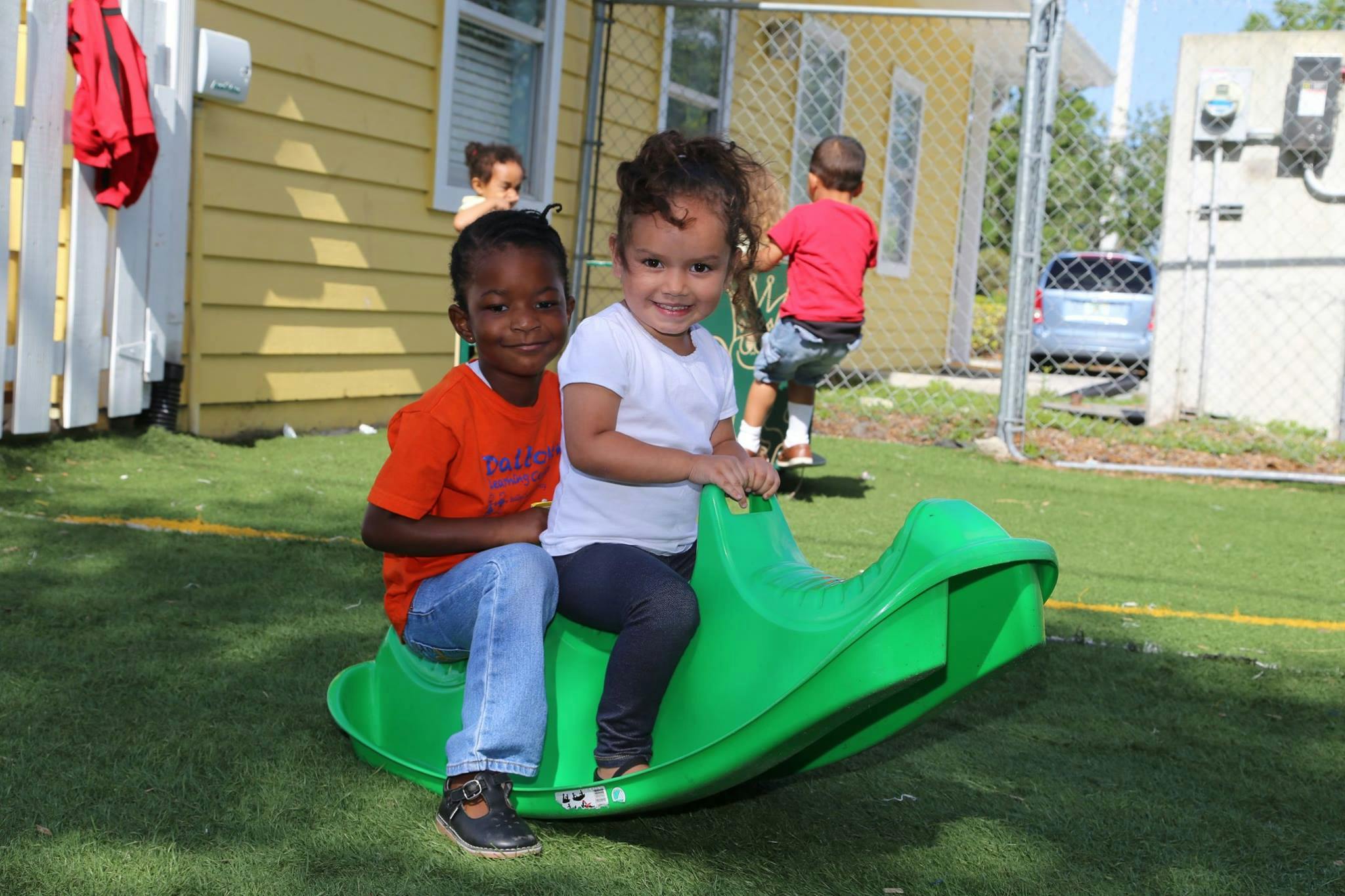 Two children on a green rocking toy, another child climbing a fence