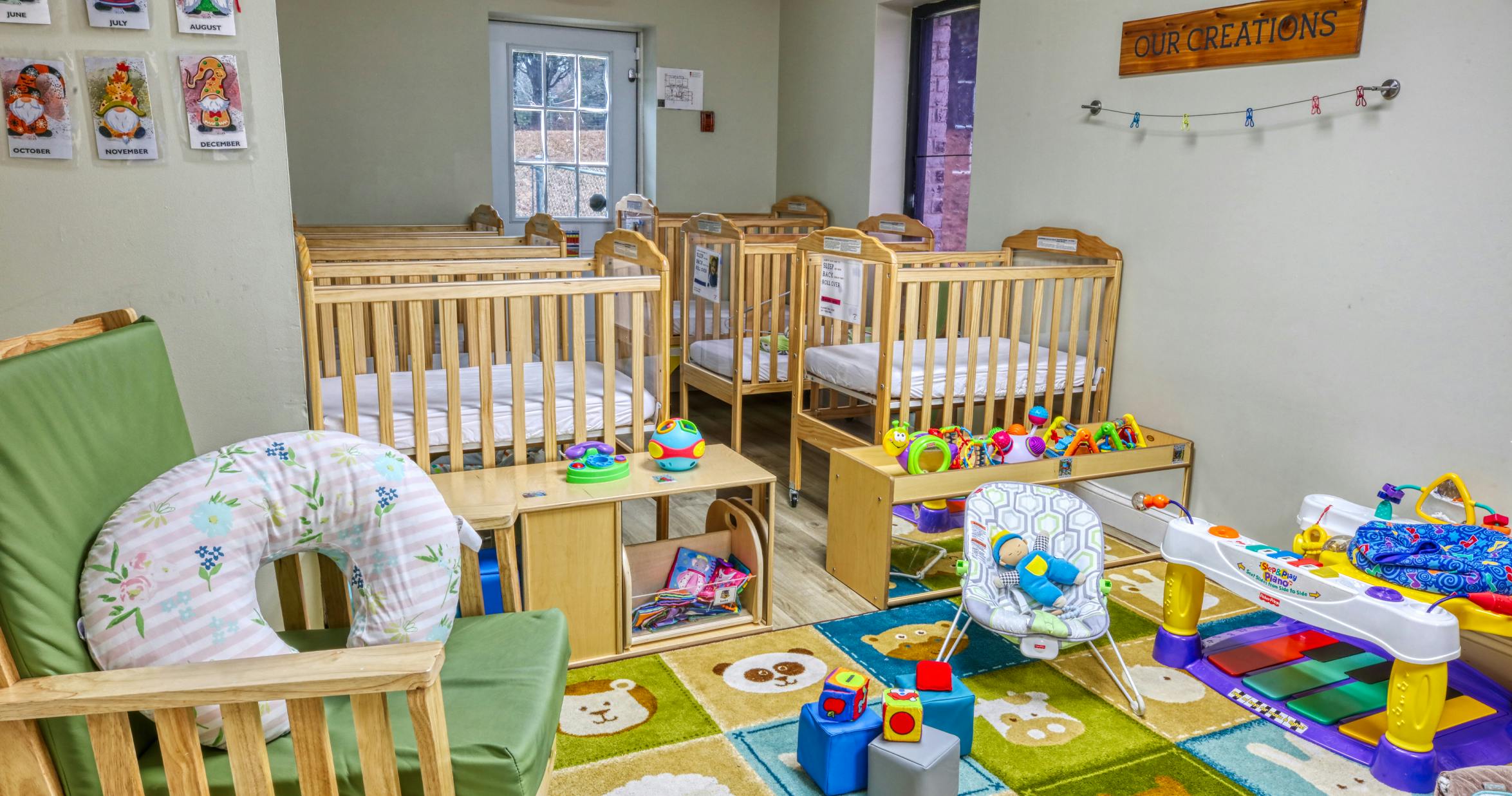 Cribs, infant seats, and colorful toys in a childcare room with a sign reading "OUR CREATIONS"