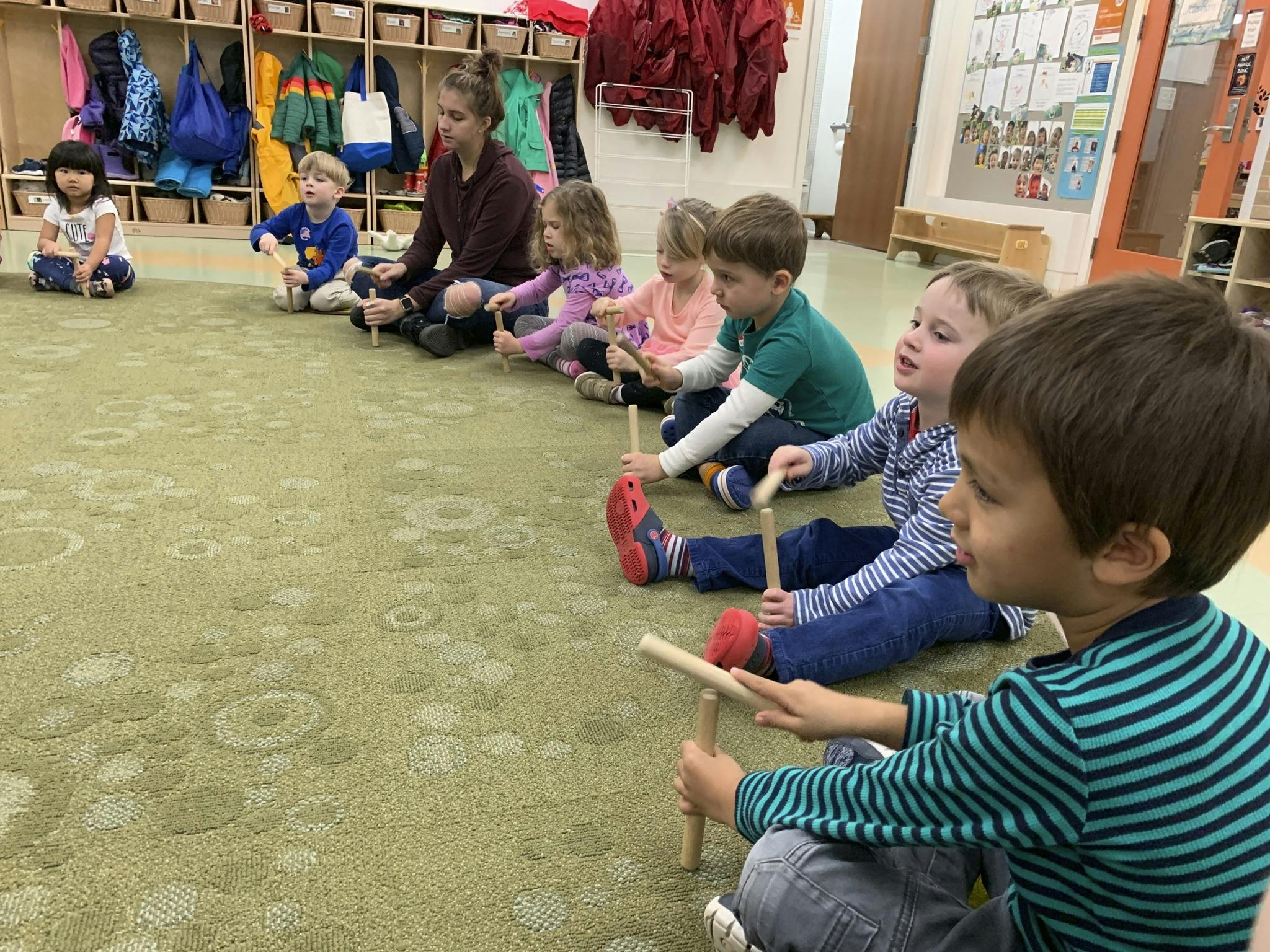 Adult and children sitting on floor holding wooden sticks in a classroom