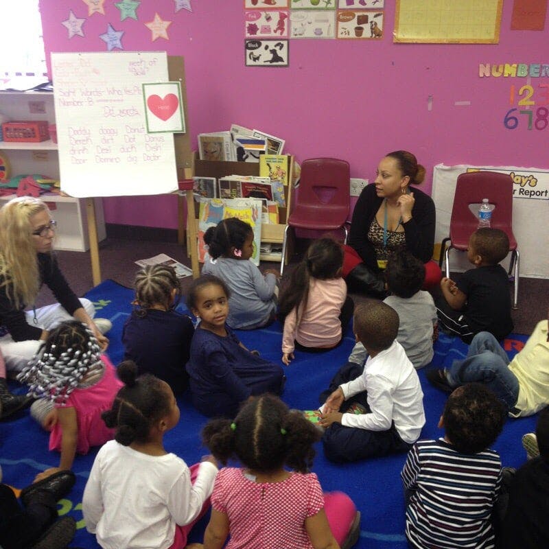 Two people sitting on the floor with a group of children in a classroom