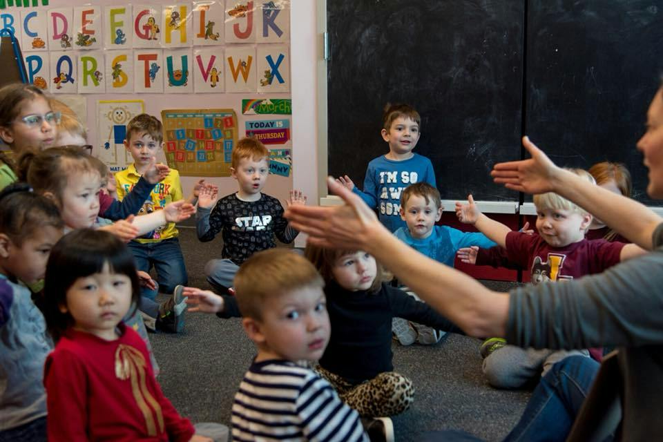 Group of children in a classroom reaching their arms out toward a person