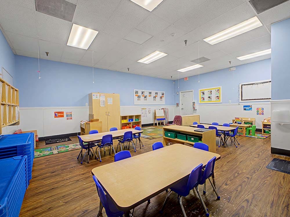 Classroom with child-sized tables and chairs, wood-patterned floor, and light blue walls