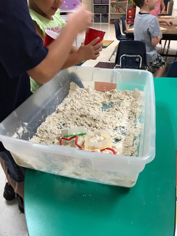 Child holding red cups over a bin of sand with cookie cutters
