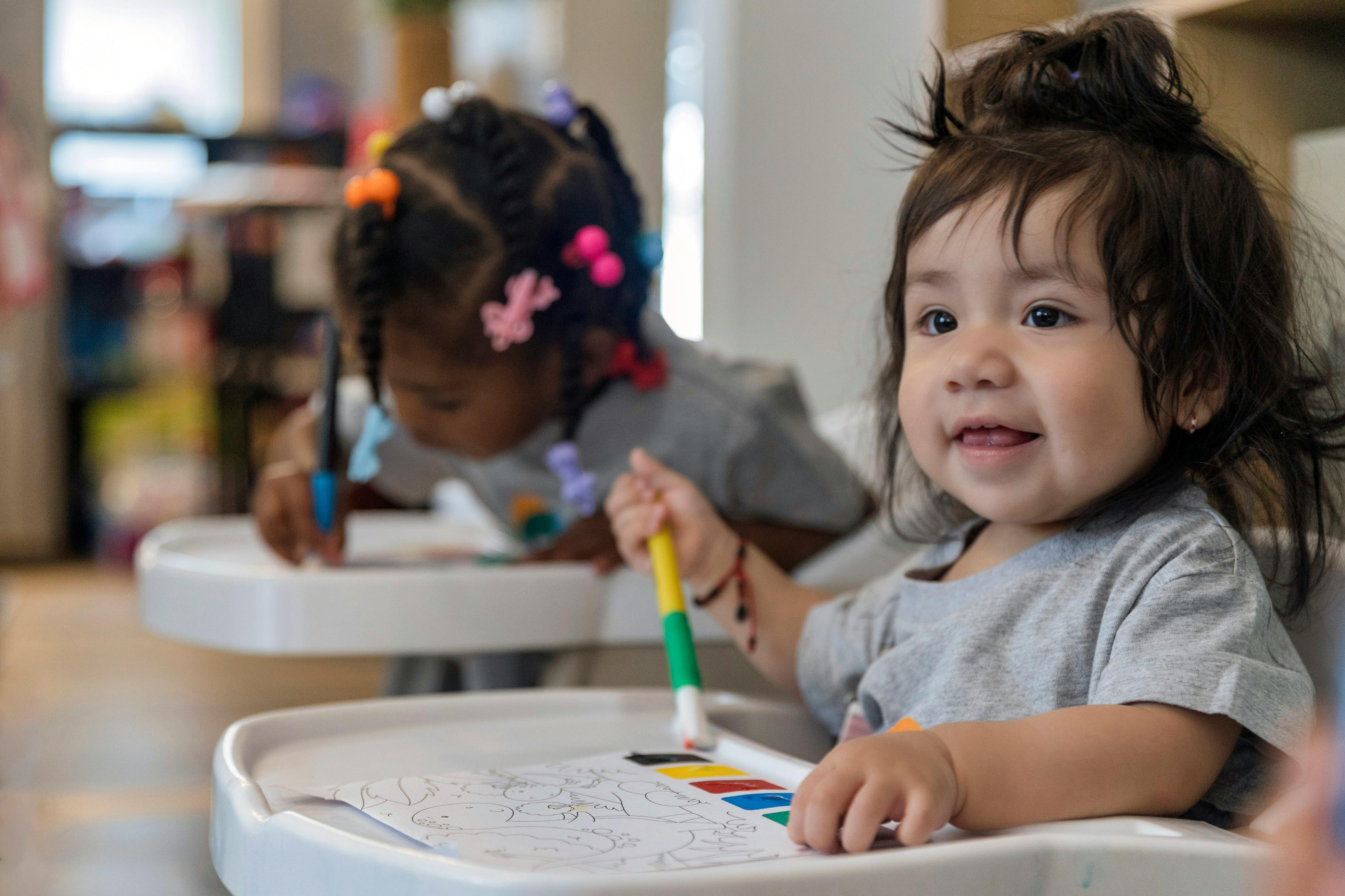 Two children in high chairs painting with one painting and the other looking forward