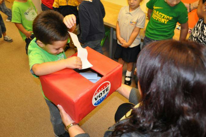 Child putting a paper into a red box with a "VOTE" sticker