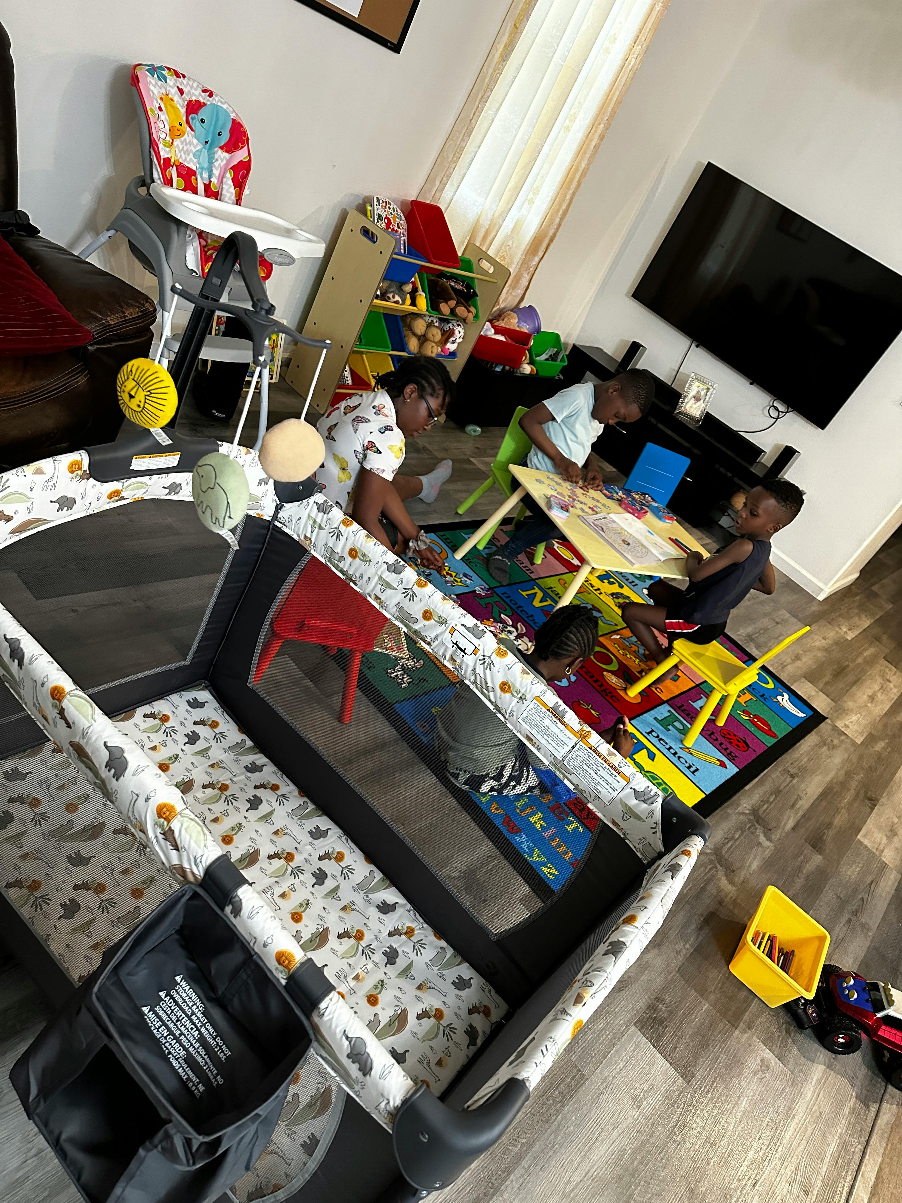 Person and two children playing at a table on a rug, with a playpen in the foreground