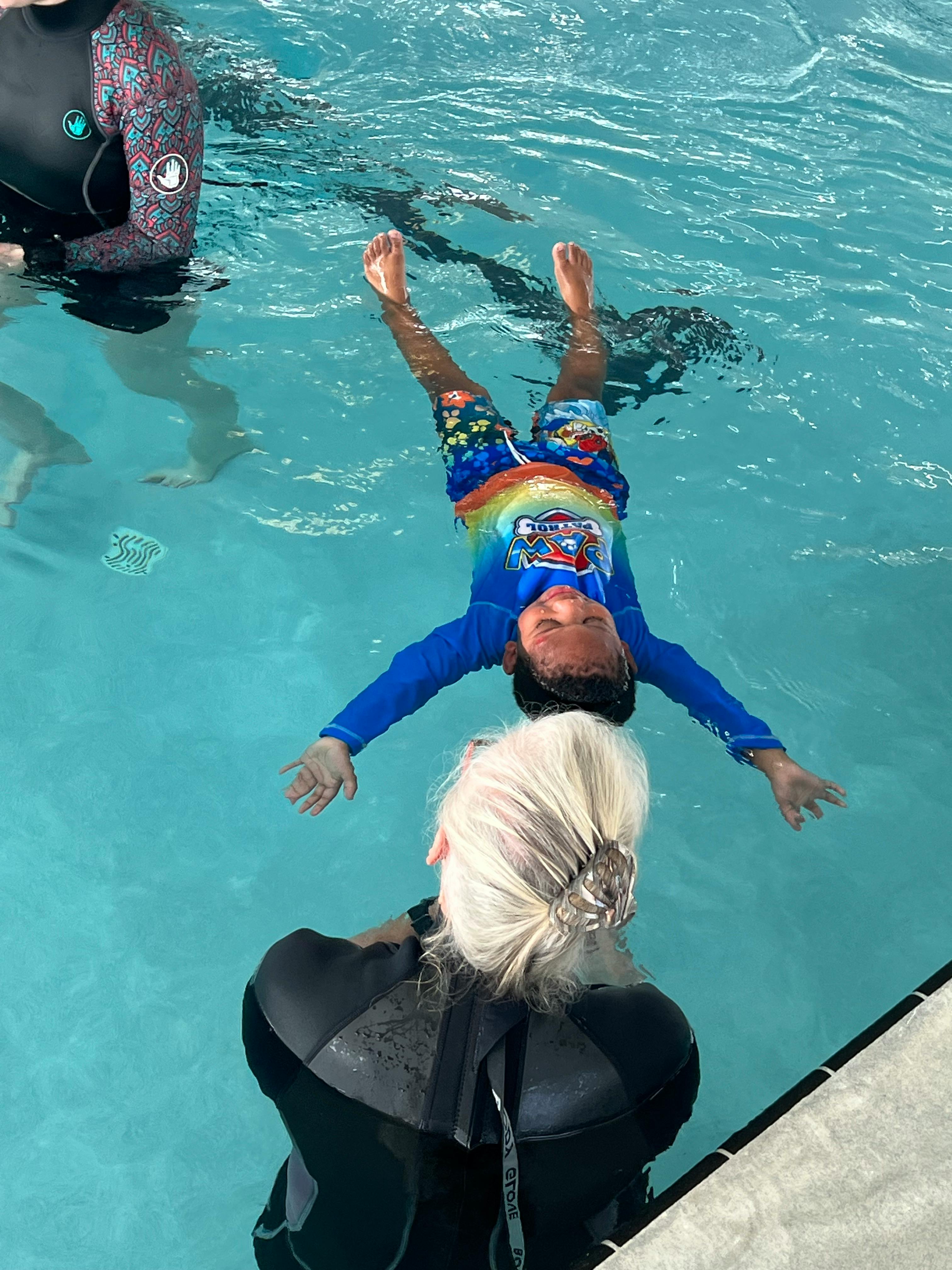 Child floating on their back in a pool with an adult nearby