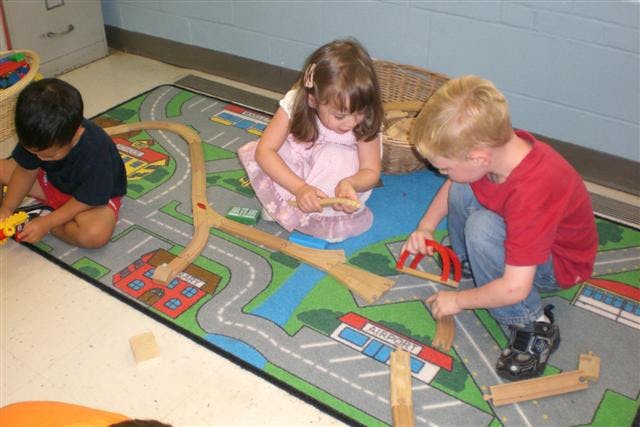 Three children playing with wooden train tracks on a rug