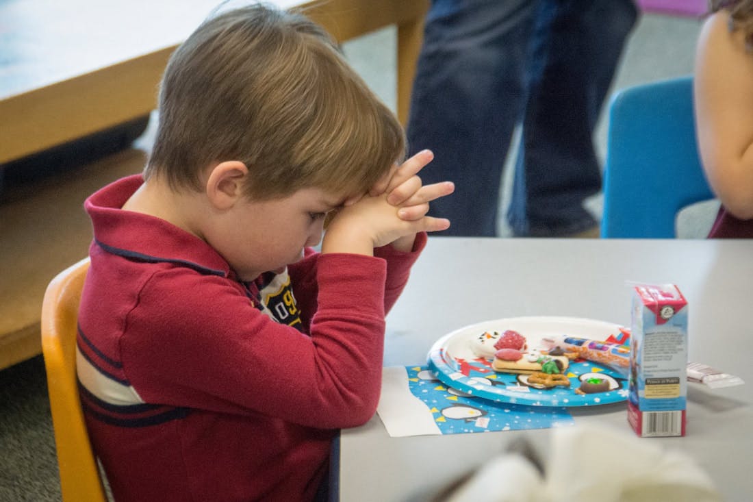 Child sitting at a table with hands clasped in front of face, next to a plate of food