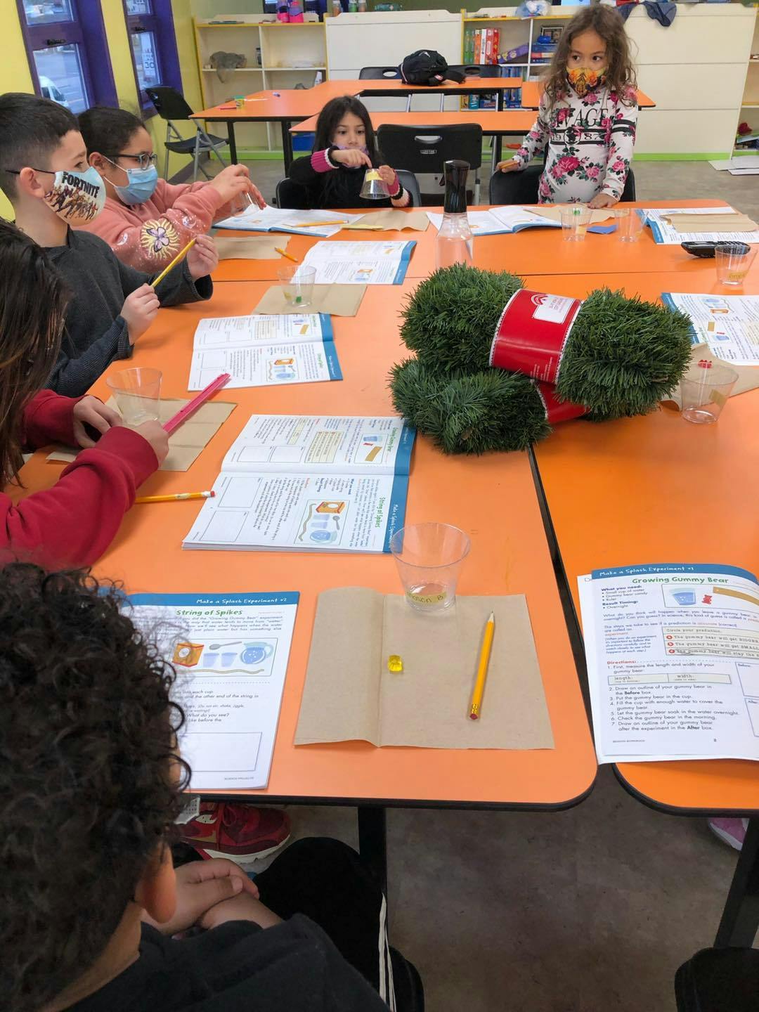 Children at desks in a classroom, some wearing masks, looking at papers