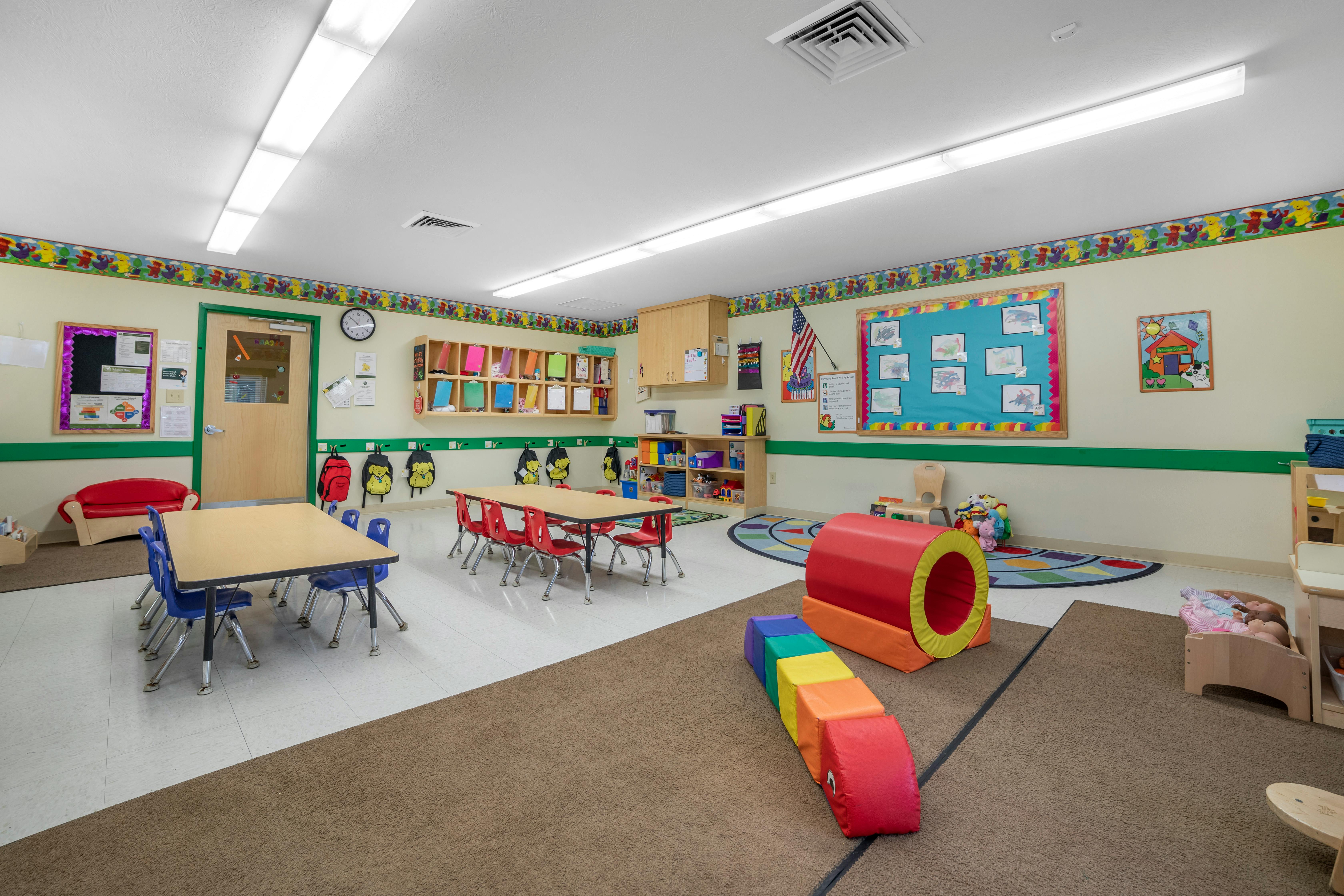 An empty childcare classroom with small tables and chairs, play equipment, and cubbies