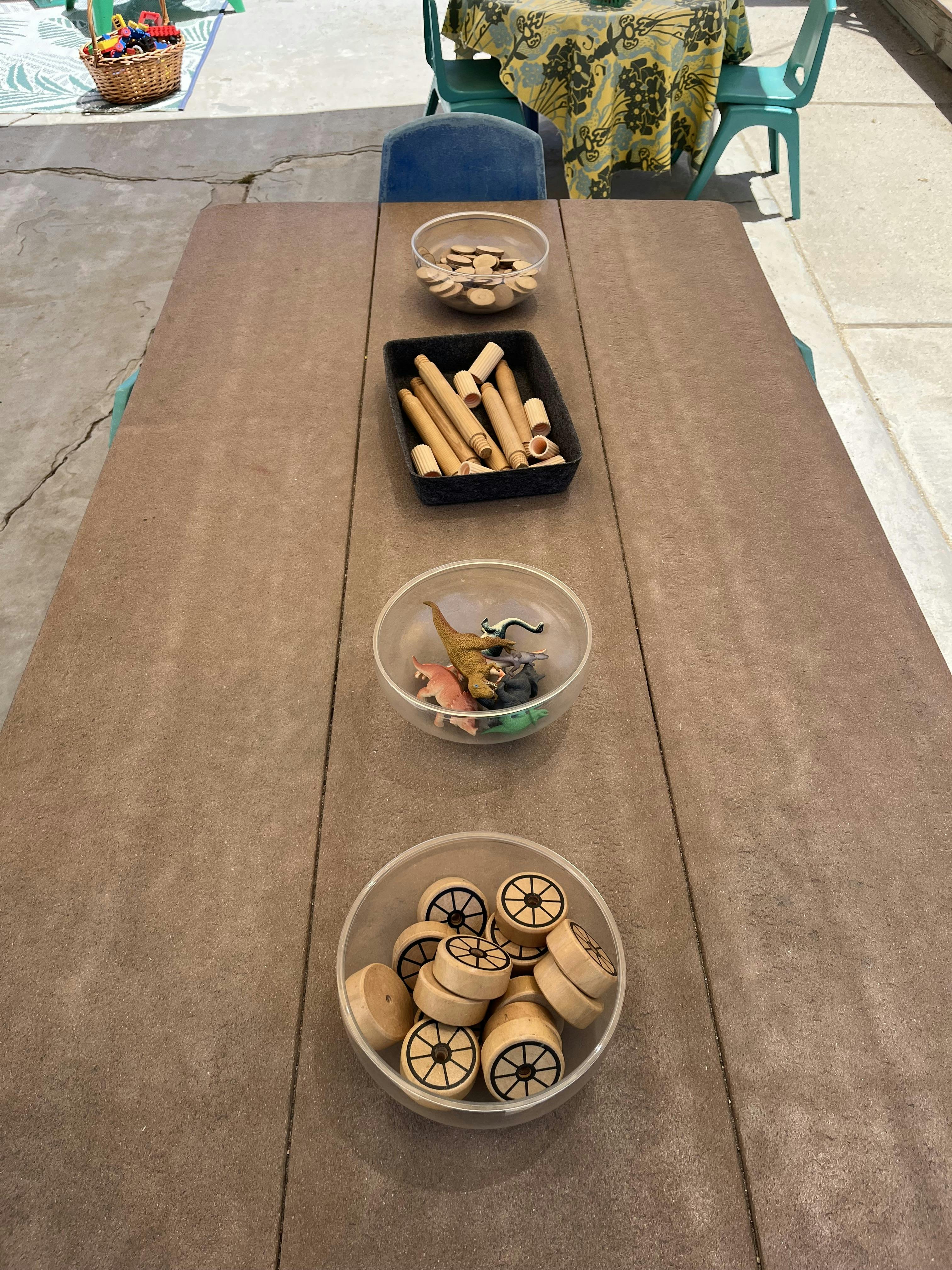 A table with bowls of wooden toy pieces, dowels, and dinosaur figurines