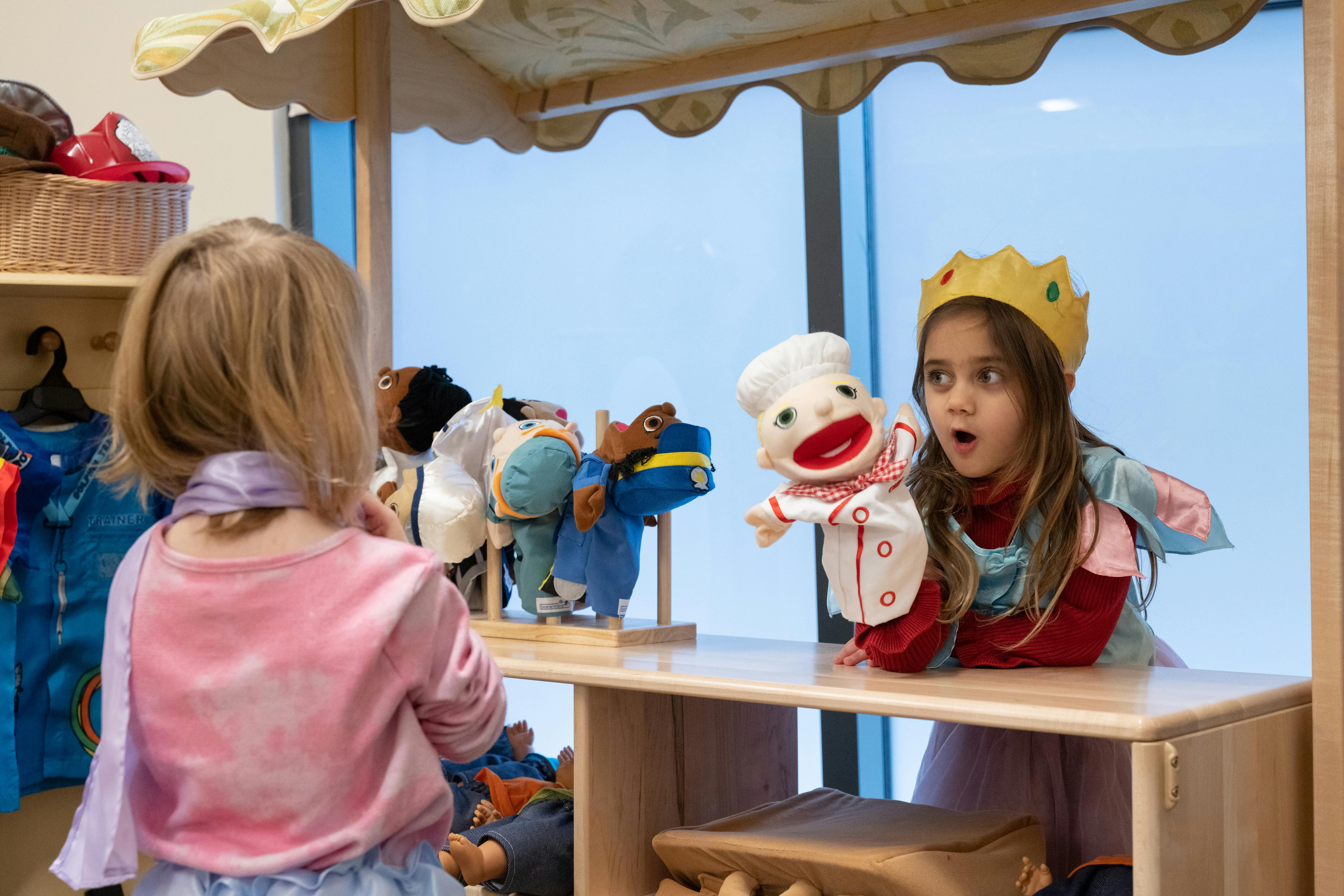 Child in crown with puppet at a puppet stand, another child watching