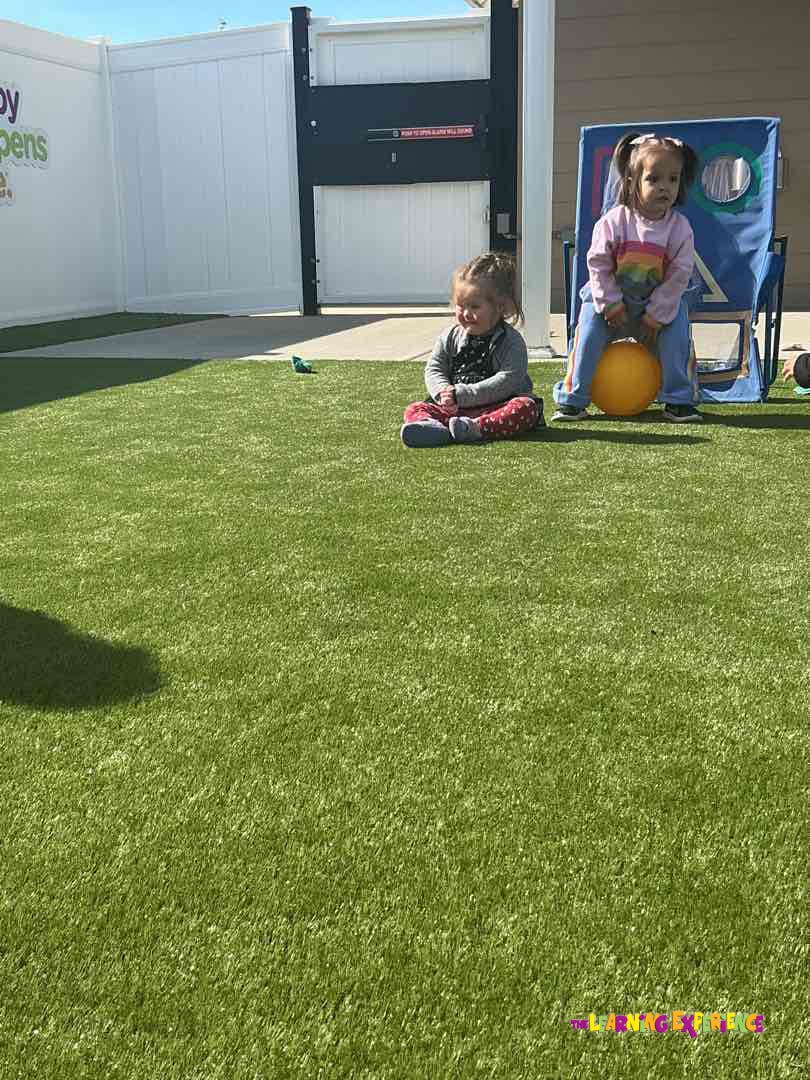 Child sitting on green turf, another child sitting on a yellow ball