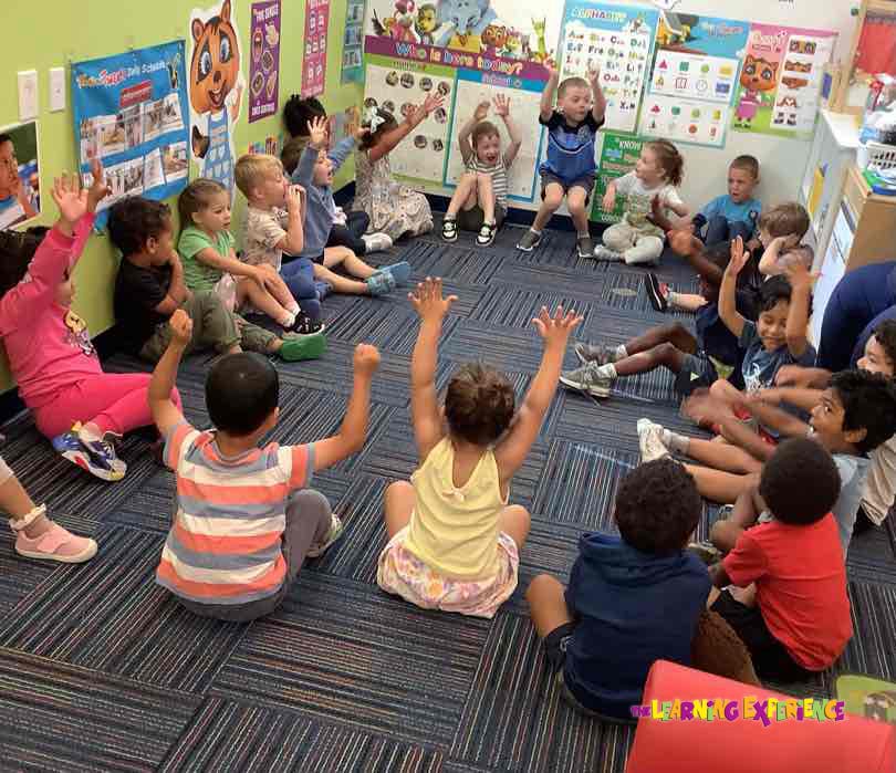 Children sitting in a circle with hands raised in a classroom