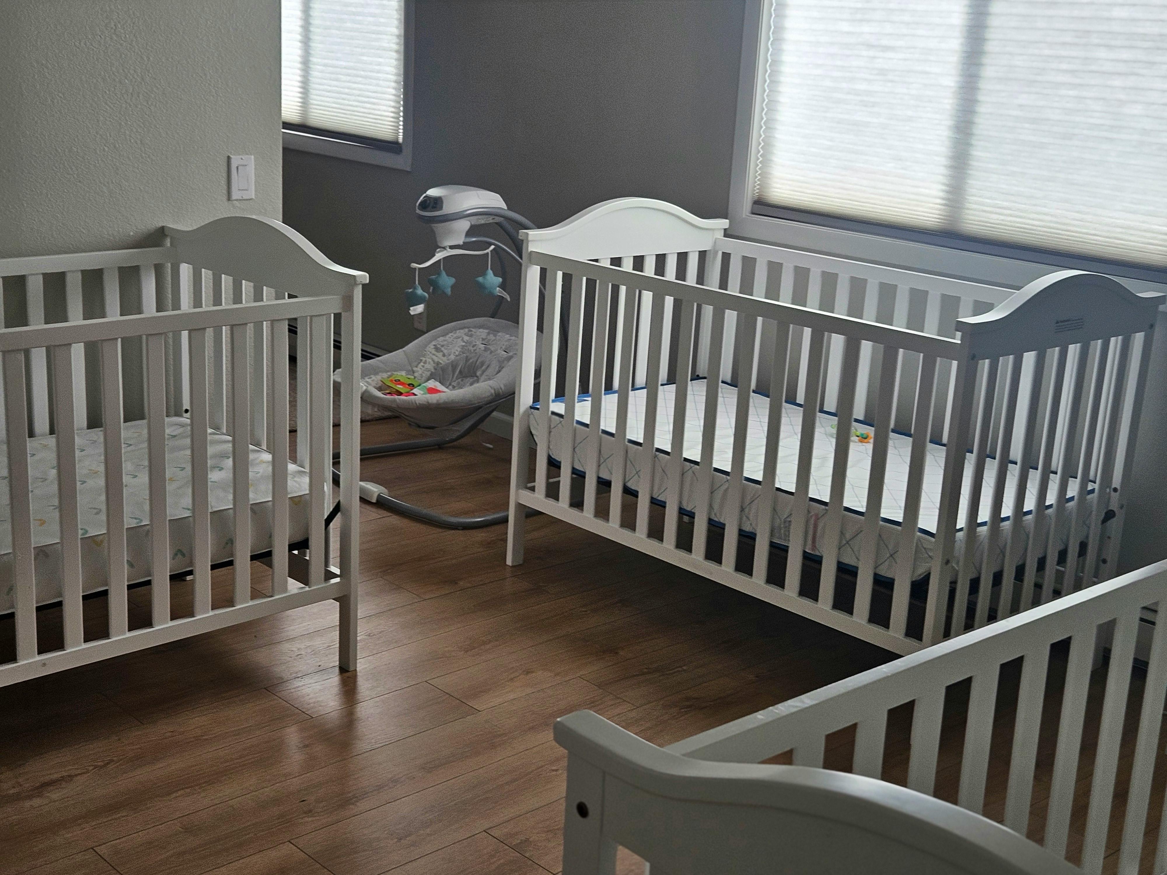 Multiple white wooden cribs and a baby bouncer in a nursery