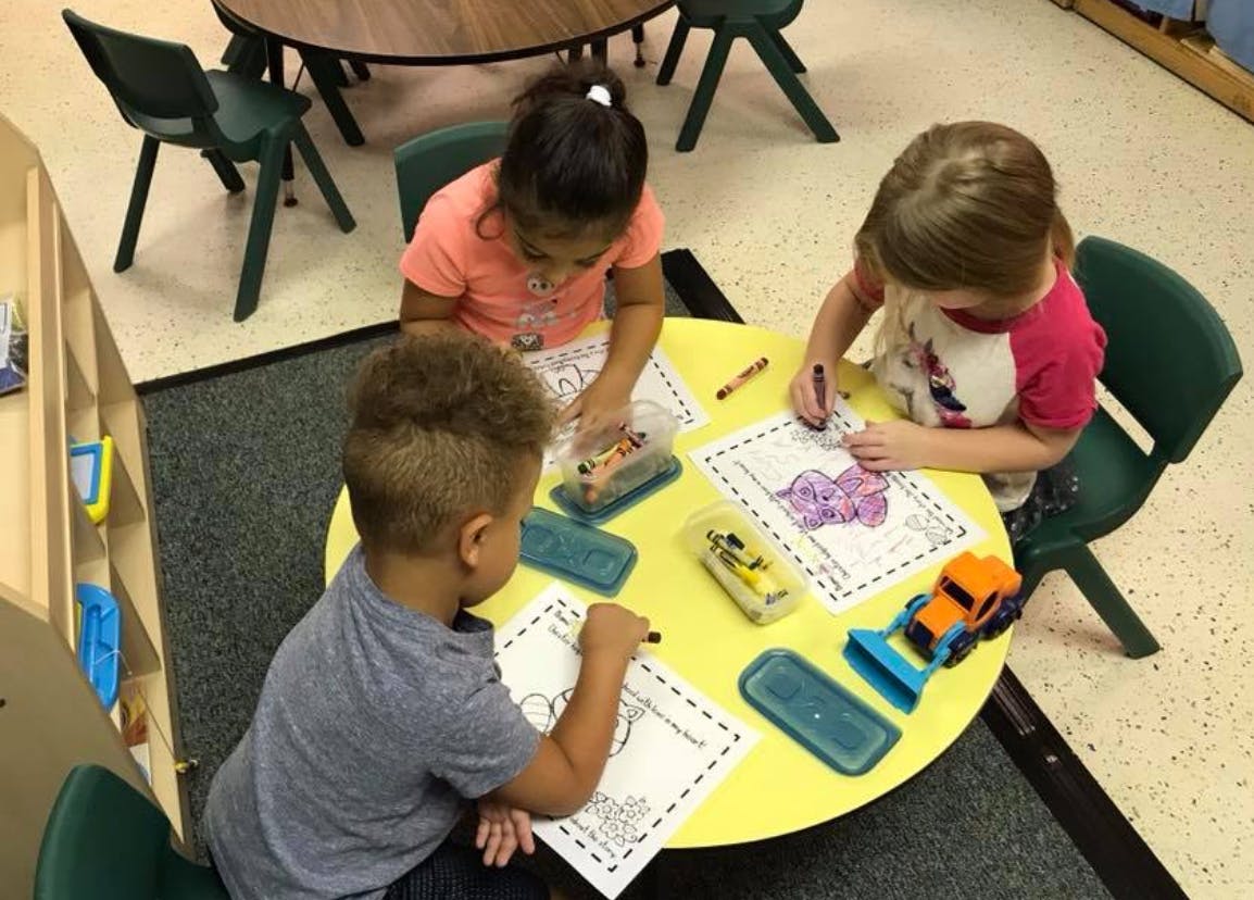 Three children coloring at a yellow table in a classroom