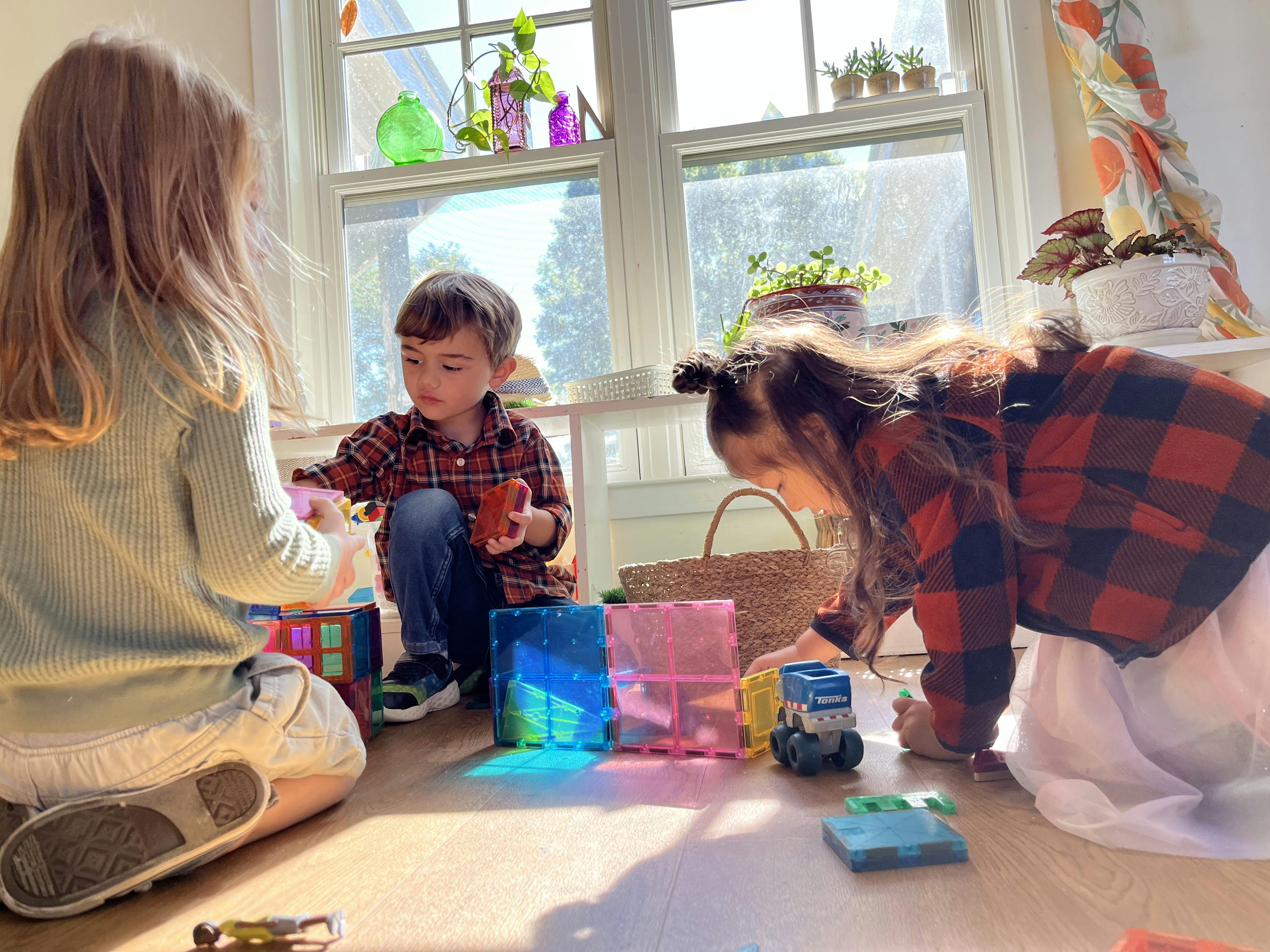 Three children playing with magnetic blocks on a wooden floor in a sunlit room