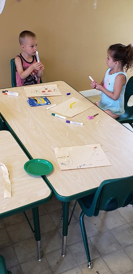 Two children sitting at a table with markers and drawings