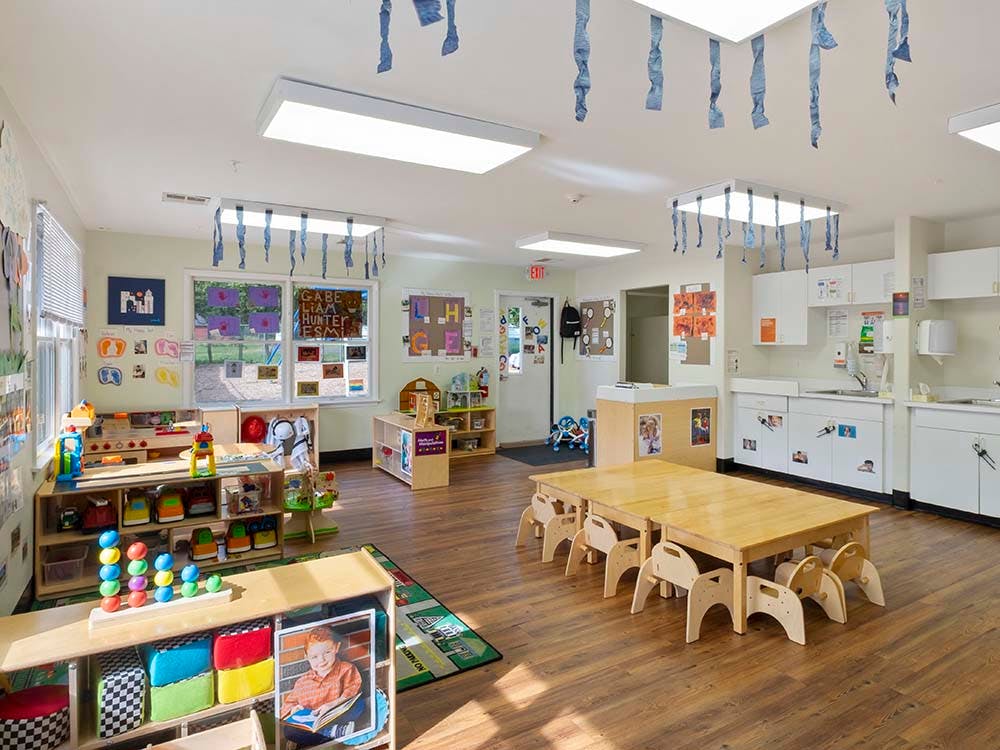 A colorful classroom with small tables and chairs, shelves with toys, and decorated windows