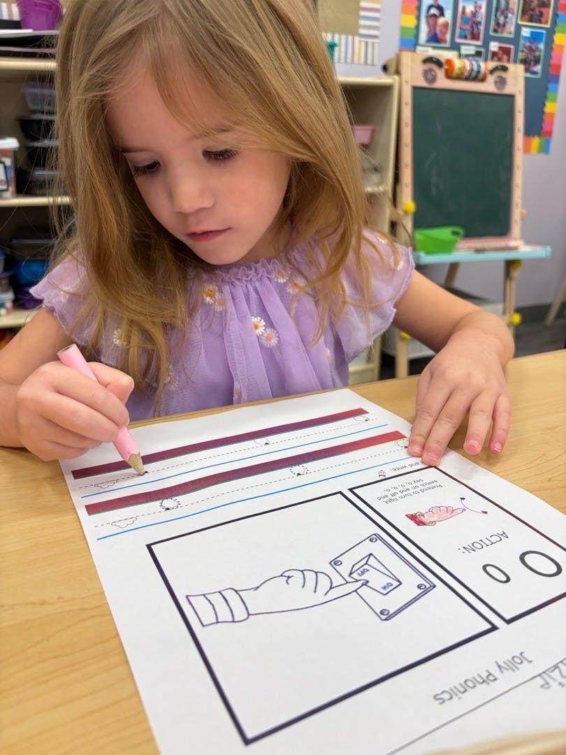 Child writing with a pink pencil on a Jolly Phonics worksheet