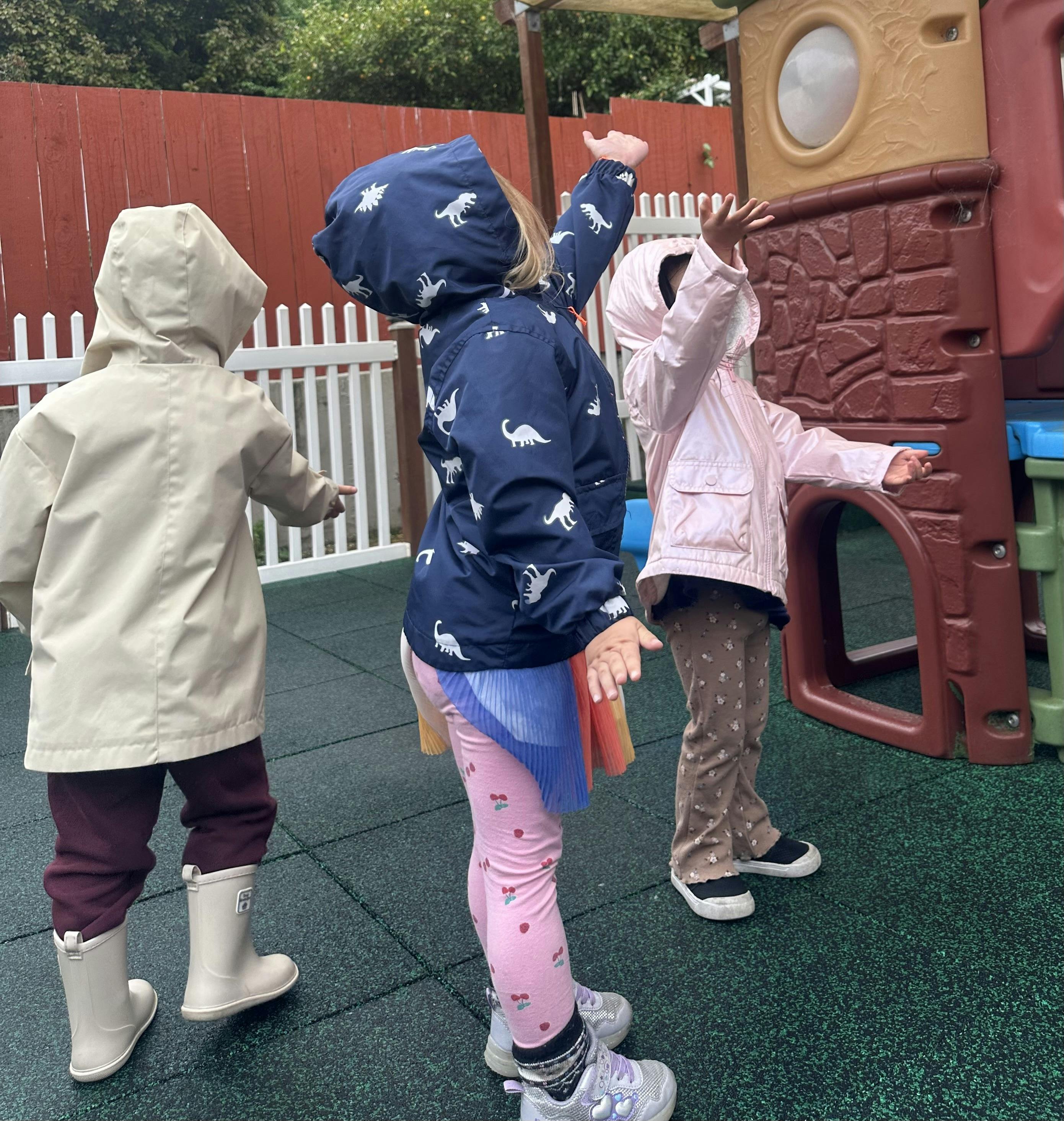 Three children in raincoats standing on a playground with arms outstretched