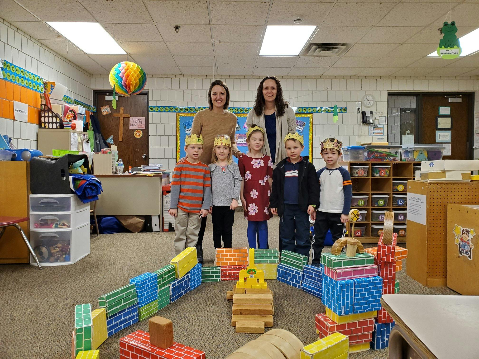 Two people and four children in a classroom with blocks on the floor