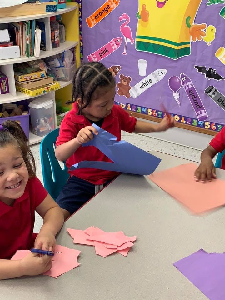 Two children cutting and folding paper at a table in a classroom