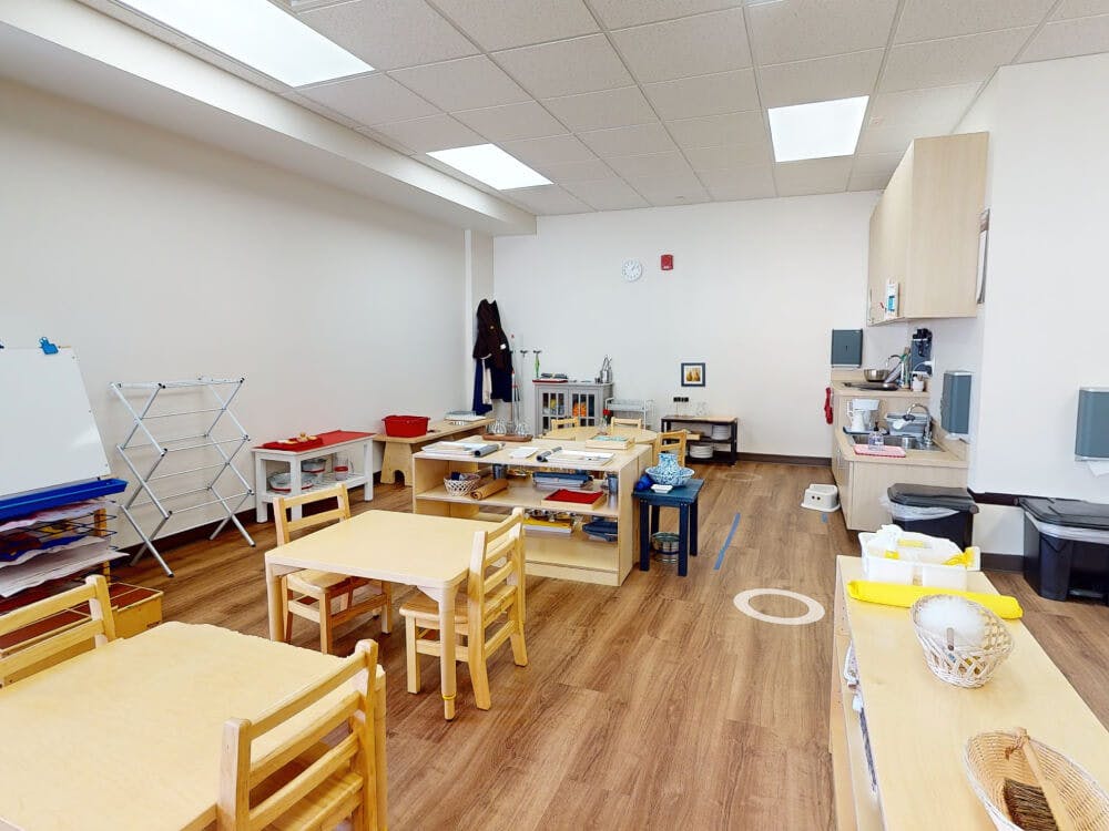An empty classroom with child-sized tables and chairs, and a small kitchen area