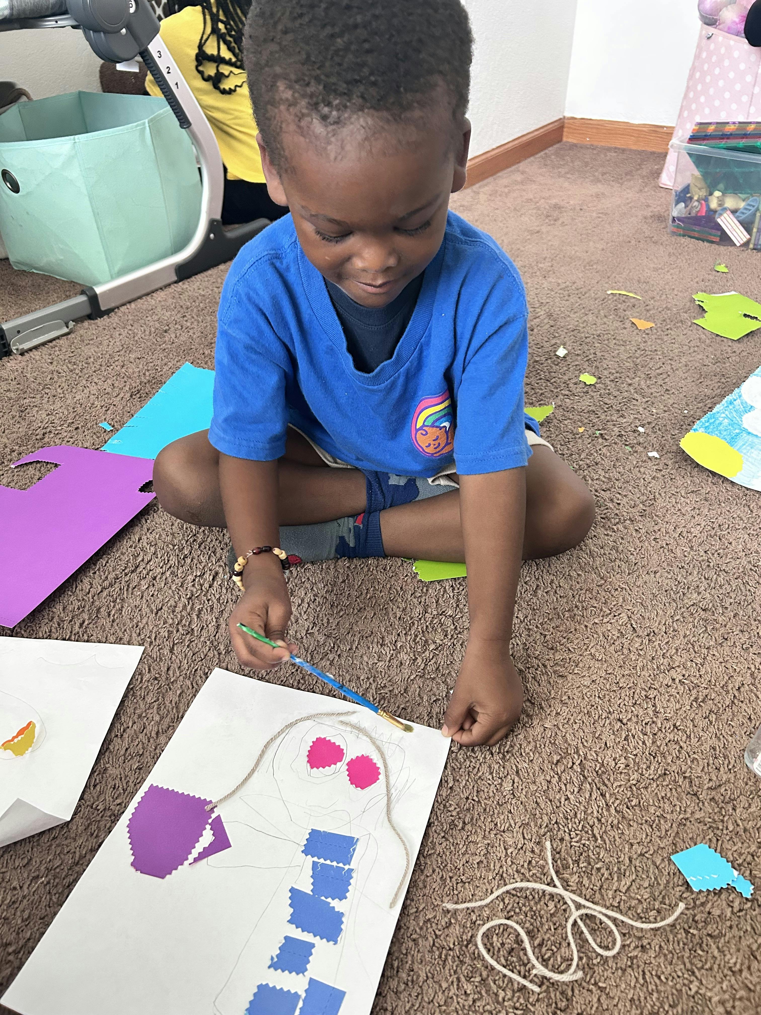 Child sitting on floor creating art with paper cutouts, yarn, and paintbrush