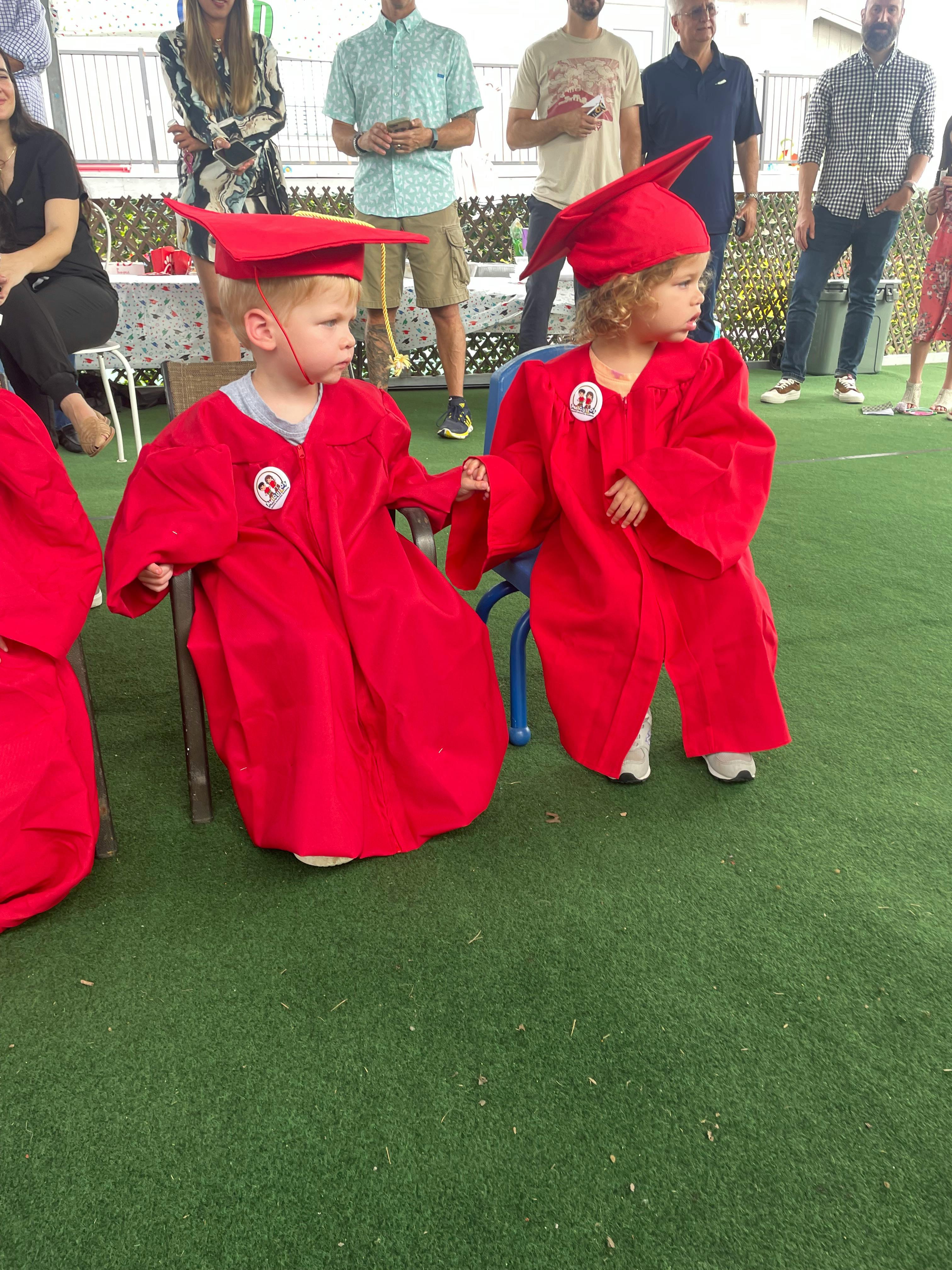 Two children in red graduation caps and gowns sitting in chairs