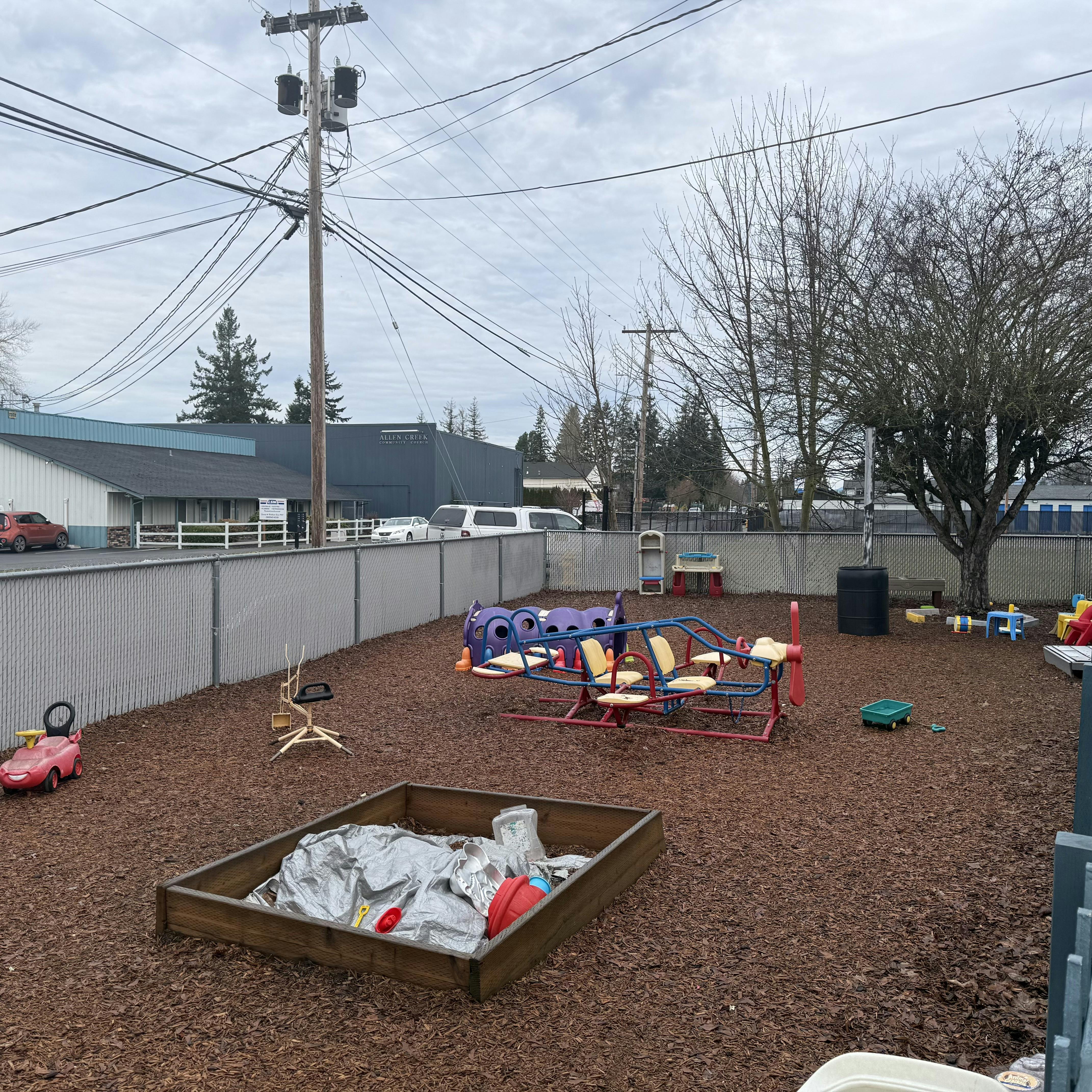 Children's playground with bark ground covering, various toys, and chain-link fence